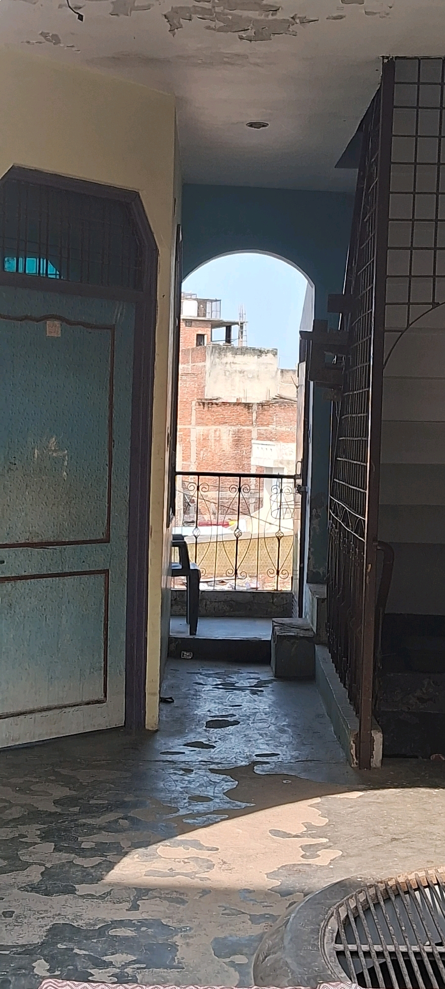 The image shows a view through an arched doorway in what appears to be a residential building in Meerut, India. The doorway leads to a balcony or outdoor space overlooking other buildings. To the left is a light blue door with dark trim and a metal grille. The floor in the foreground is concrete with some areas of water or dampness, reflecting sunlight. A metal grate, possibly a drain, is visible in the lower right corner.
The outdoor space beyond the arch features a decorative metal railing and a chair. The buildings visible outside are constructed of brick and concrete, with some appearing to be unfinished or under construction. The sky is clear and blue. The overall impression is of an urban environment with modest architecture. The scene is static, with no people or overt actions depicted. The lighting suggests it is daytime, likely morning or afternoon, given the angle of the shadows.