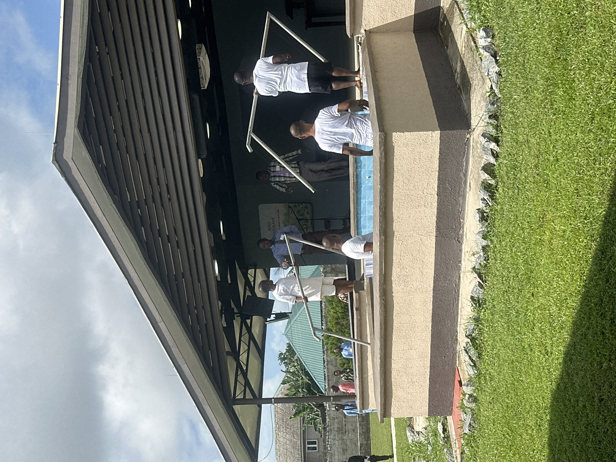 In Itoga, Nigeria, outdoors, on a bright day with scattered clouds, a baptism is taking place in a raised, tiled pool. The pool's exterior is a light tan with a dark brown band around its base. A group of men are present.
In the pool, a man wearing a white t-shirt and black shorts stands with his back to the camera. In front of him, a man in a white robe and shorts is partially submerged. To his right, another man in a white t-shirt and black shorts is standing, facing away from the camera and holding a white frame. Another white frame is being held by a man further back, who is wearing a white t-shirt and black shorts, facing away from the camera.
Behind the pool, on a raised platform, two men are talking. One is wearing a light blue shirt and dark pants, and the other is in a checkered shirt and grey pants. Behind them, a large green sign with text in white and green is visible.
To the left of the pool, several other men are standing. One man in a white t-shirt and light shorts stands on the edge of the pool. Another man in a light blue shirt and black pants stands behind him. Further to the left, a man in a dark suit stands on the grass.
In the background, a building with a brown, slatted roof extends over the platform. Houses with green roofs and a tall tree are visible behind the building. The area around the pool is covered in lush green grass with scattered grey rocks.