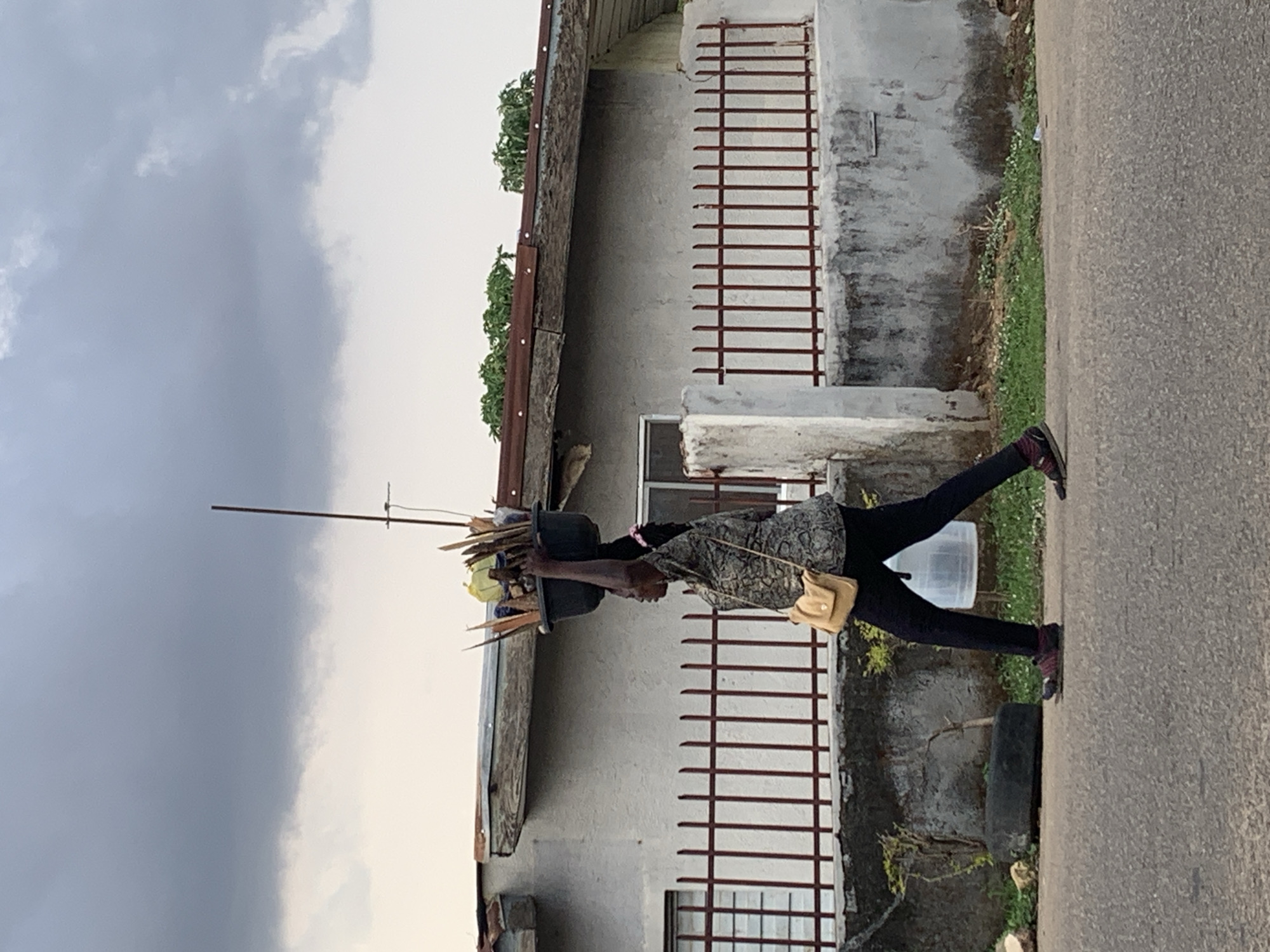 A woman carries items on her head while walking along a road in Jos, Nigeria. She is wearing black leggings, a patterned blouse, and carrying a small tan purse. The items on her head include black pots, sticks, and other objects balanced precariously. She is walking in front of a building with barred windows and a rusty metal roof. The sky is overcast. The road is gray asphalt.