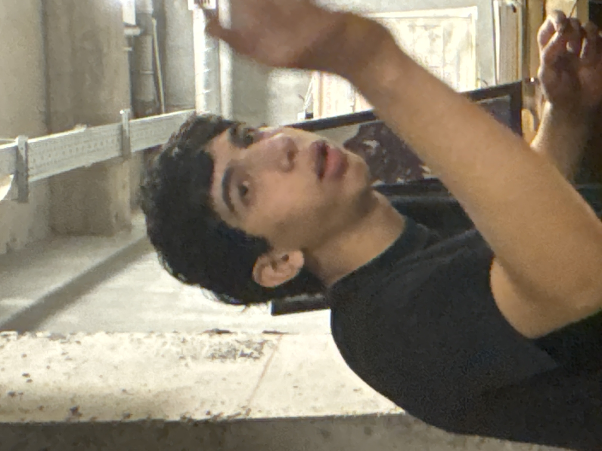 A young man with dark hair and olive skin is visible in a close-up, low-angle shot. He is wearing a dark, possibly black, T-shirt. His right arm is extended upwards and partially out of frame, while his left arm is closer to his body. The man's head is tilted back, and he is looking upwards with his mouth slightly open.
The background features industrial-looking elements, including pipes and metal supports. There is also a ledge or shelf with a textured surface in the foreground.
The location cannot be determined to be Yerevan, Armenia, with the available visual information. There are no distinguishing landmarks or features to confirm this.