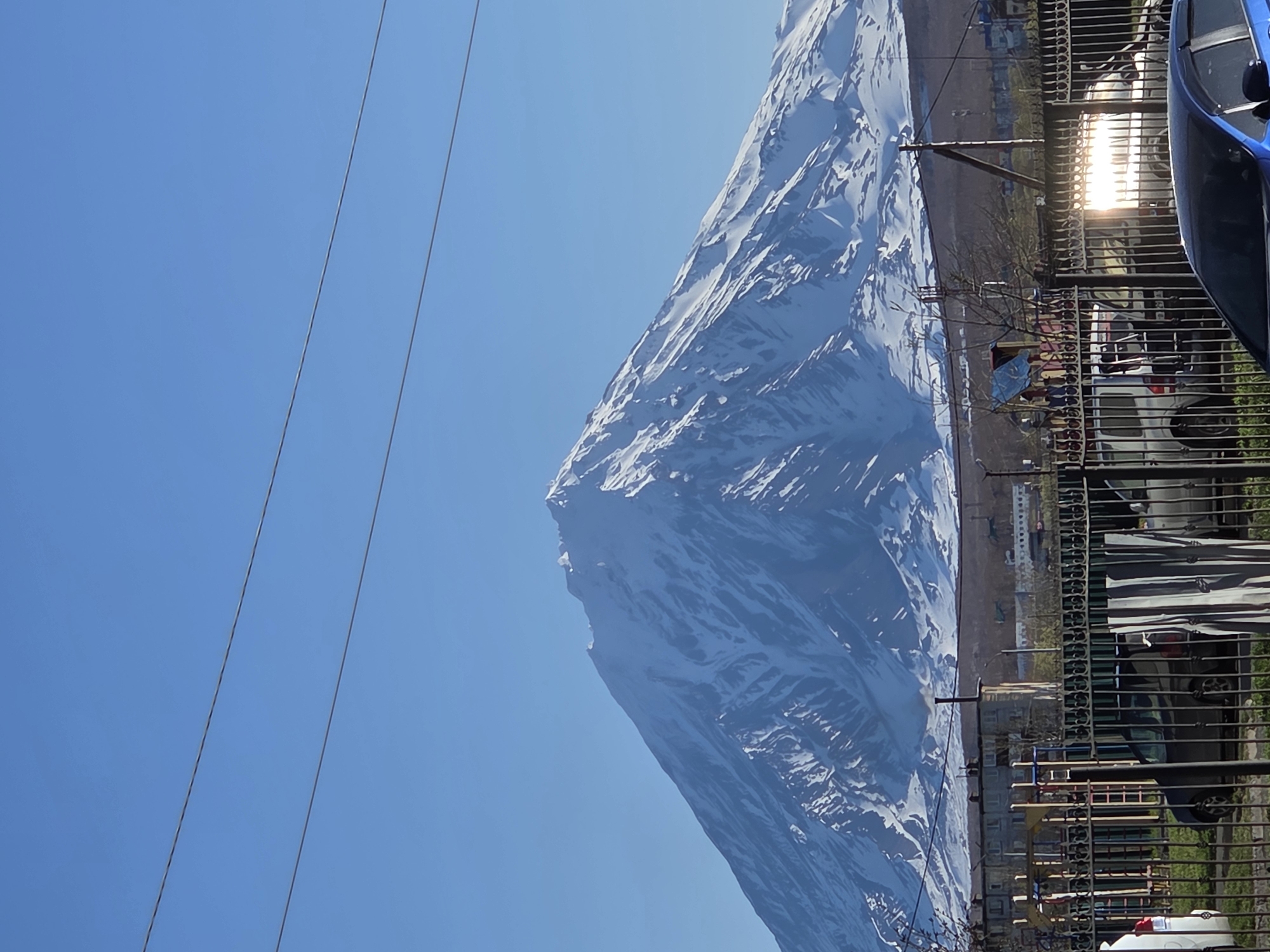 The image depicts a large, snow-covered mountain rising above the city of Petropavlovsk-Kamchatsky, Russia. The sky is clear and blue. In the foreground, a metal fence and parked cars partially obscure the view. Power lines are visible crossing the sky. The city buildings appear to be located at the base of the mountain. Sunlight reflects off the fence.