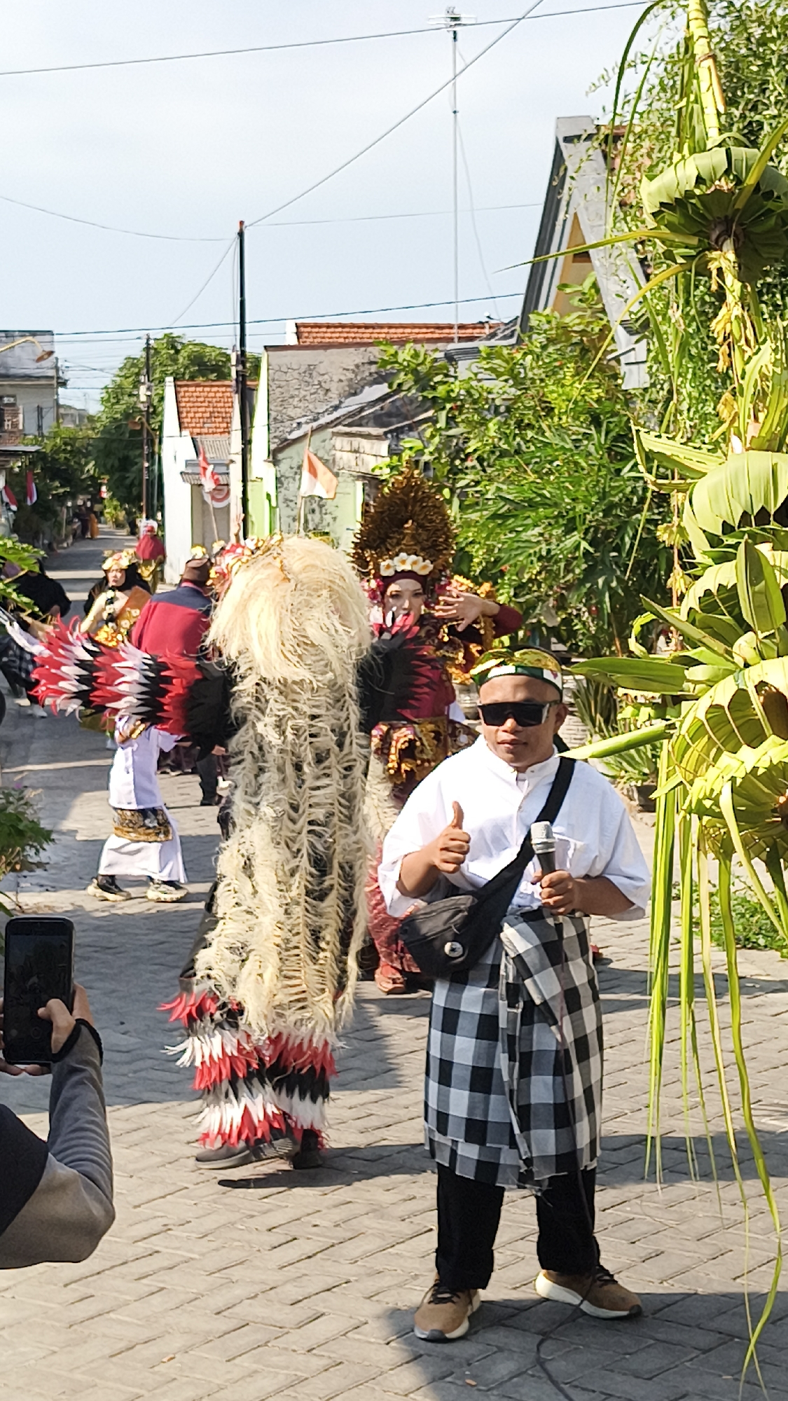 The image depicts a street scene in Dalegan, Indonesia, featuring a procession or performance. In the foreground, a man wearing a white shirt, black pants, and a checkered sarong over his pants gives a thumbs-up gesture while holding a microphone. He is also wearing sunglasses and a green and yellow patterned cap. Behind him, a person in a large, elaborate costume with flowing white and red fringe is visible. Further back, other individuals in traditional attire, some with headdresses and colorful feathered costumes, are part of the procession. Indonesian flags are displayed on some of the buildings lining the street, suggesting a festive or national occasion. The environment includes paved walkways, buildings with red-tiled roofs, and lush green foliage. A person on the left side of the frame is holding up a smartphone, seemingly to record the event.