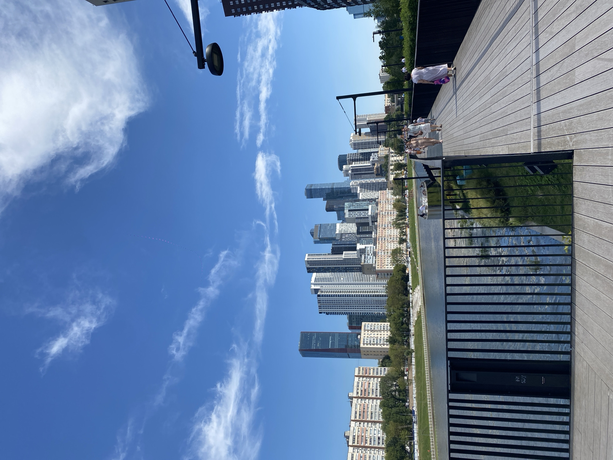 This image depicts a sunny day in Moscow, Russia, likely at the Moscow River. In the foreground, a wooden pedestrian walkway runs diagonally from the bottom right towards the left, with several people strolling along it. A black metal railing lines the walkway. Beyond the railing, the Moscow River is visible, with a dark, vertical grate structure in the water near the foreground.
In the background, a cityscape is dominated by numerous tall, modern buildings, indicative of a business or residential district. These skyscrapers are of varying heights and architectural styles, including a prominent dark-red-toned skyscraper. Between the river and the buildings, there is a green embankment with trees and some lower-rise residential buildings. The sky is a vibrant blue, dotted with white, wispy clouds. There are also tiny, dark specks visible high in the sky that could be birds. A modern street lamp with a black fixture is visible on a black pole extending from the top right of the frame. The overall scene suggests a pleasant, urban outdoor environment.