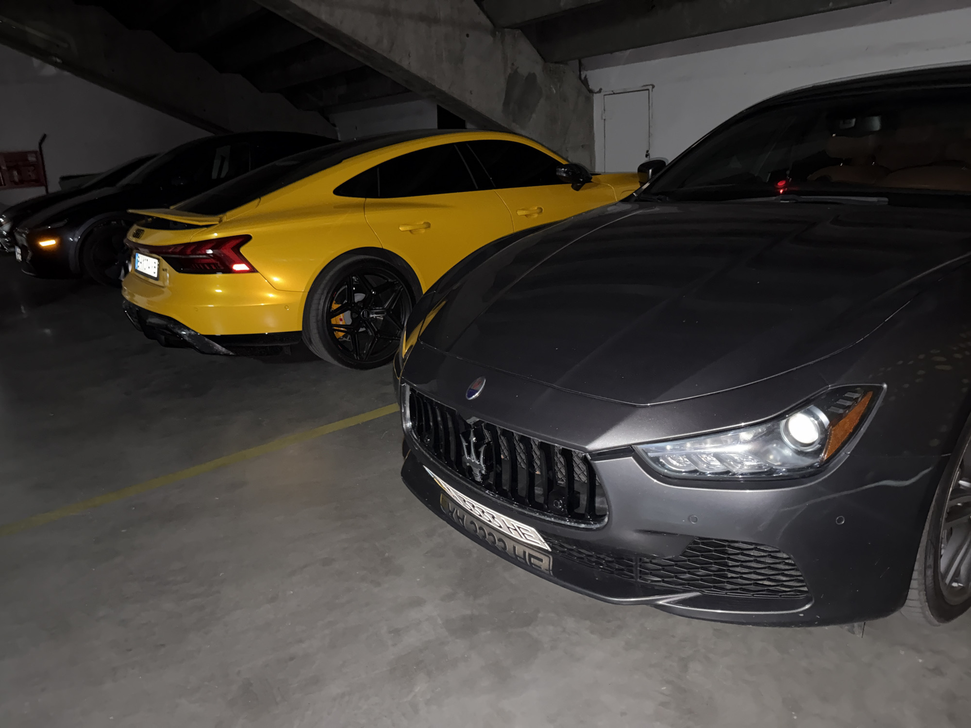 The image shows three cars parked indoors, likely in a garage. In the foreground, a dark gray Maserati Ghibli is prominently displayed, its front grille and headlights clearly visible. The Maserati has a license plate with Cyrillic characters. Behind it, a bright yellow Audi RS e-tron GT is parked, showing its sleek design and black wheels. Further in the background, a dark-colored car, possibly another sedan or coupe, is partially visible.
The scene is dimly lit, suggesting it is nighttime or the garage has limited lighting. The environment appears to be a concrete structure with exposed beams overhead, typical of a parking garage. A yellow line is painted on the concrete floor, marking a parking space. The overall impression is one of luxury and automotive display, with the high-performance vehicles taking center stage. There is no indication of people or any specific activity taking place beyond the cars being parked. The location context of Odesa, Ukraine, is noted.