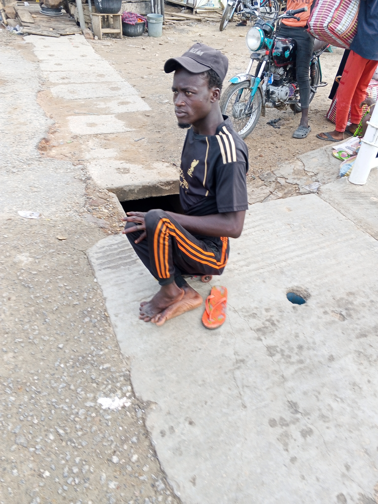 A man in Zango-ndaji, Nigeria squats on a concrete slab next to a sewer grate. He wears a dark baseball cap, black short-sleeved shirt with gold stripes on the sleeves and the Liverpool football club logo on the chest, and black pants with orange stripes down the legs. He is barefoot, with an orange flip-flop beside his feet. In the background are motorcycles, other people, and debris.