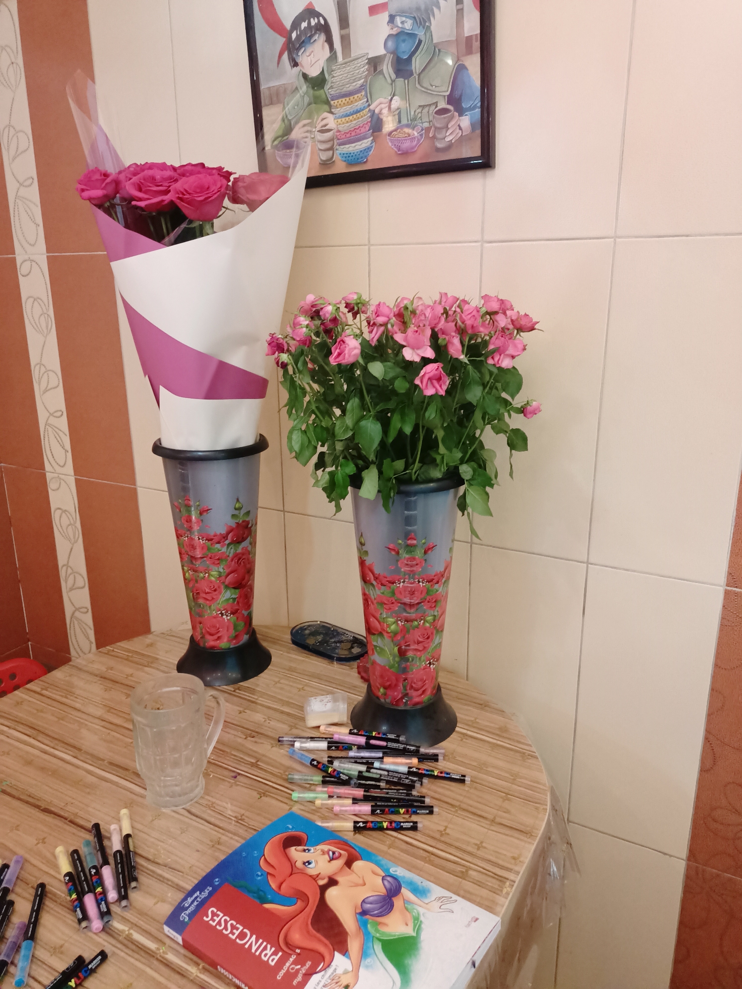 The image displays a still life scene on a wooden table, with a tiled wall in the background. In the upper portion of the image, a framed picture depicts two animated characters. To the left and right of the framed picture, two vases hold bouquets of pink roses. The vase on the left contains a larger bouquet, while the vase on the right holds a smaller, more compact arrangement.
On the table, a clear glass mug is present. A coloring book featuring Disney princesses, specifically Ariel and another mermaid, lies open. Scattered around the coloring book and near the mug are numerous markers, predominantly in black, pink, blue, and yellow, suggesting an art or craft activity. The overall impression is of a domestic setting, likely a kitchen or dining area, given the tiled walls and countertop. There is no discernible information in the image to suggest a location in Almaty, Kazakhstan.