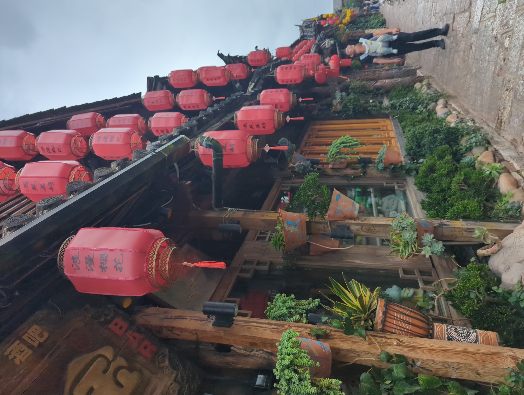 In Gucheng District, China, a street scene unfolds with traditional architecture and festive decorations. The buildings are adorned with numerous red lanterns, some featuring Chinese characters. Lush green plants cascade from terracotta pots on wooden beams and along the stone pathway. A person, likely a parent, is visible in the lower right, wearing a baby carrier with a child. They are looking upwards, possibly at the lanterns. The atmosphere suggests a culturally rich and visually appealing urban environment.