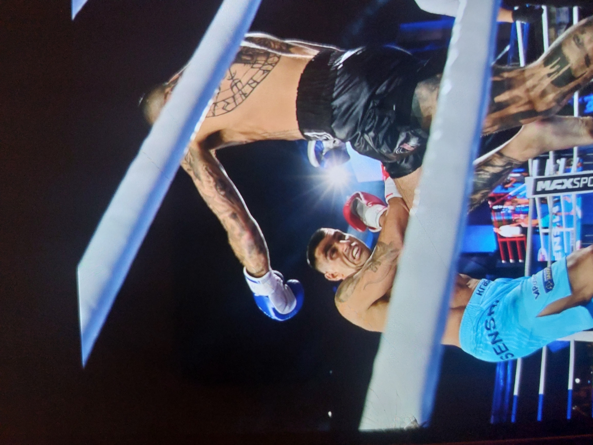 An action-packed photograph captures two male boxers engaged in a match within a boxing ring. The image is rotated, presenting the scene from an unusual angle.
One fighter, positioned towards the upper-left of the frame, wears black boxing shorts with a faint Adidas logo and blue boxing gloves. He is heavily tattooed, with intricate designs covering his arms, a large geometric pattern across his back/side, and tattoos extending down his legs. He appears to be leaning backward, potentially evading or reacting to an opponent's move.
The second fighter, visible in the lower-right, wears light blue boxing shorts and red boxing gloves. He also has tattoos adorning his arms. His face is clearly visible, turned upwards with an intense, focused expression.
The setting is a boxing ring, demarcated by two bright white ropes that traverse the image. The background is dark, suggesting an indoor arena during an evening event, with strong spotlights illuminating the fighters and casting reflections.
Visible text includes "SENSORY" and Cyrillic characters "КГВ" on the light blue shorts (inverted due to the image's rotation). In the background, a sign partially reads "MAXSPORT", indicating a sports channel or brand, along with other Cyrillic script. The presence of Cyrillic text supports the location context of Tsarevo, Bulgaria. The dynamic composition freezes a moment of high energy and concentration during the match.
