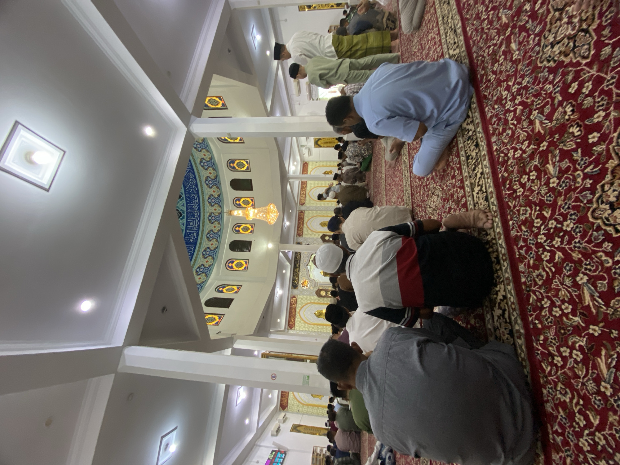 The image depicts an interior view of a mosque in the city of Lhokseumawe, Indonesia, during a prayer gathering. A large number of men are seated on a red carpet with an intricate floral pattern, facing a mihrab which is not fully visible but indicated by the arrangement of people. The architecture features high ceilings with recessed lighting and decorative elements, including arched windows with colorful stained glass and Arabic calligraphy adorning the upper walls. Several white pillars support the structure. In the foreground, three men are seated, viewed from behind. The man on the left wears a gray shirt, the central man wears a shirt with red, white, and black stripes, and the man on the right wears a light blue outfit. Further back, other individuals are visible, some standing and some seated, engaged in prayer. Three men stand near the center of the image, one wearing a white cap and traditional attire, another in a green outfit, and a third in a white shirt and green sarong. A large, ornate chandelier hangs from the ceiling above the central dome. The overall scene conveys a sense of communal religious observance.