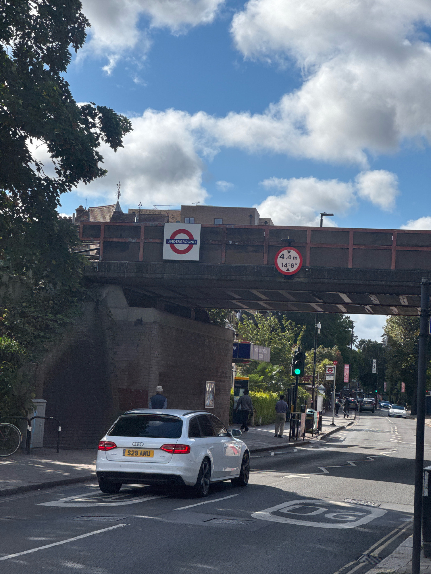 The image shows a street scene in London, United Kingdom, under a railway bridge. A white Audi A4 estate car, license plate S29 AMU, is driving on the road. Overhead, a sign for the London Underground is visible, along with a height restriction sign indicating 4.4 meters (14'6"). The sky is blue with white clouds. A traffic light is green. People are walking on the sidewalk. The environment is urban with trees and buildings visible.
