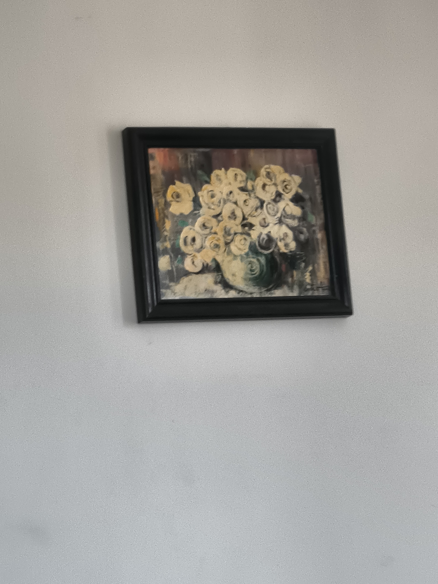 A framed painting of a floral arrangement hangs on a light gray or white wall. The frame is black. The painting depicts a cluster of white and pale yellow flowers in a green vase. The background is a mix of dark colors. The painting is positioned in the upper-center area of the image. No people or specific location details are visible.