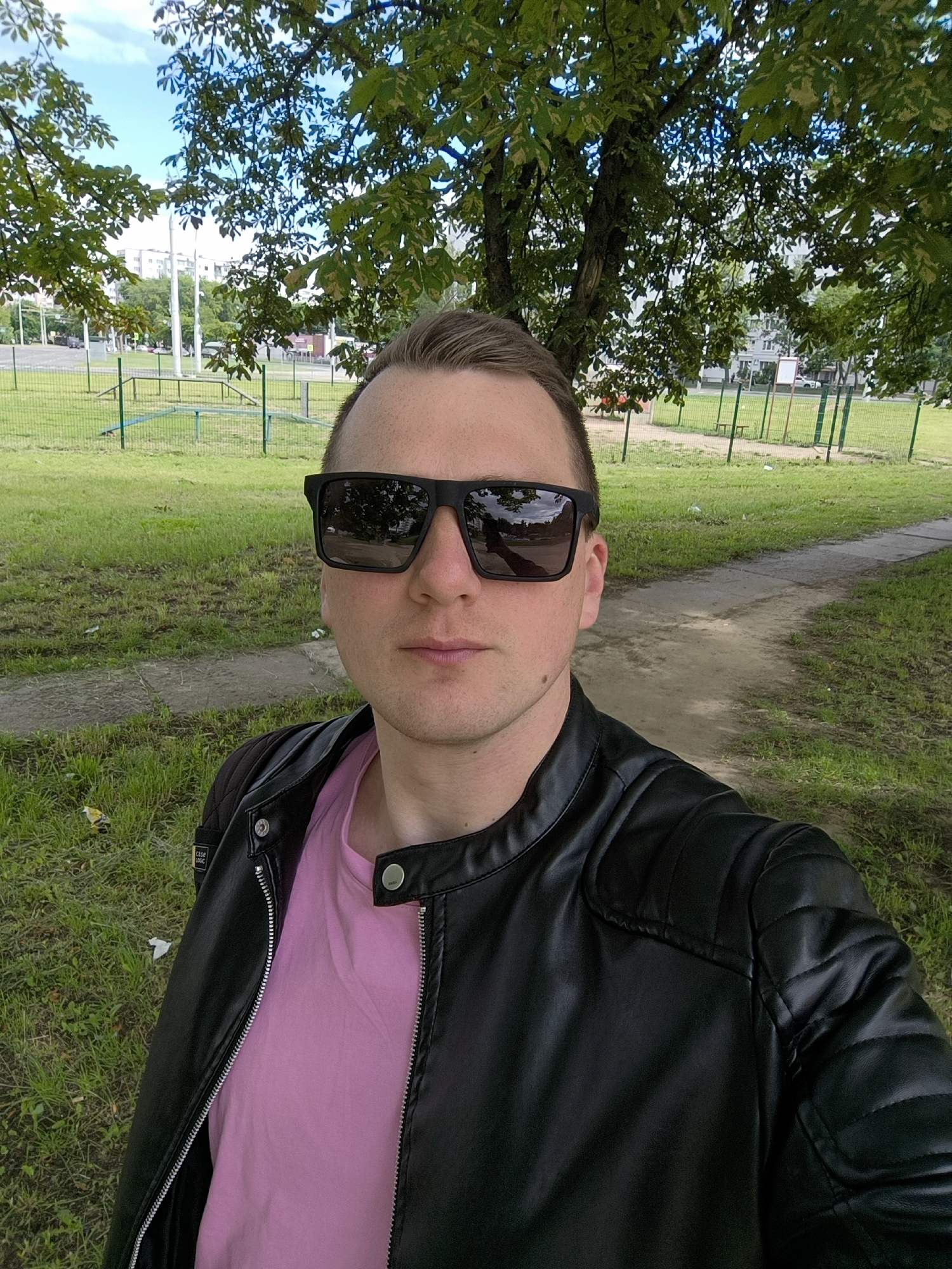 This media file is a selfie taken outdoors in Minsk, Belarus. The subject is a man wearing sunglasses, a pink t-shirt, and a black faux leather jacket. He is standing in front of a tree with green leaves, and behind him, a grassy area with a path is visible. In the background, there is a fence, some playground equipment, and apartment buildings. The sky is blue with some clouds, suggesting it is daytime and the weather is pleasant. There is some text visible on a tag on the jacket, which appears to read "CASE LOGIC". The overall impression is a casual, daytime outdoor scene.