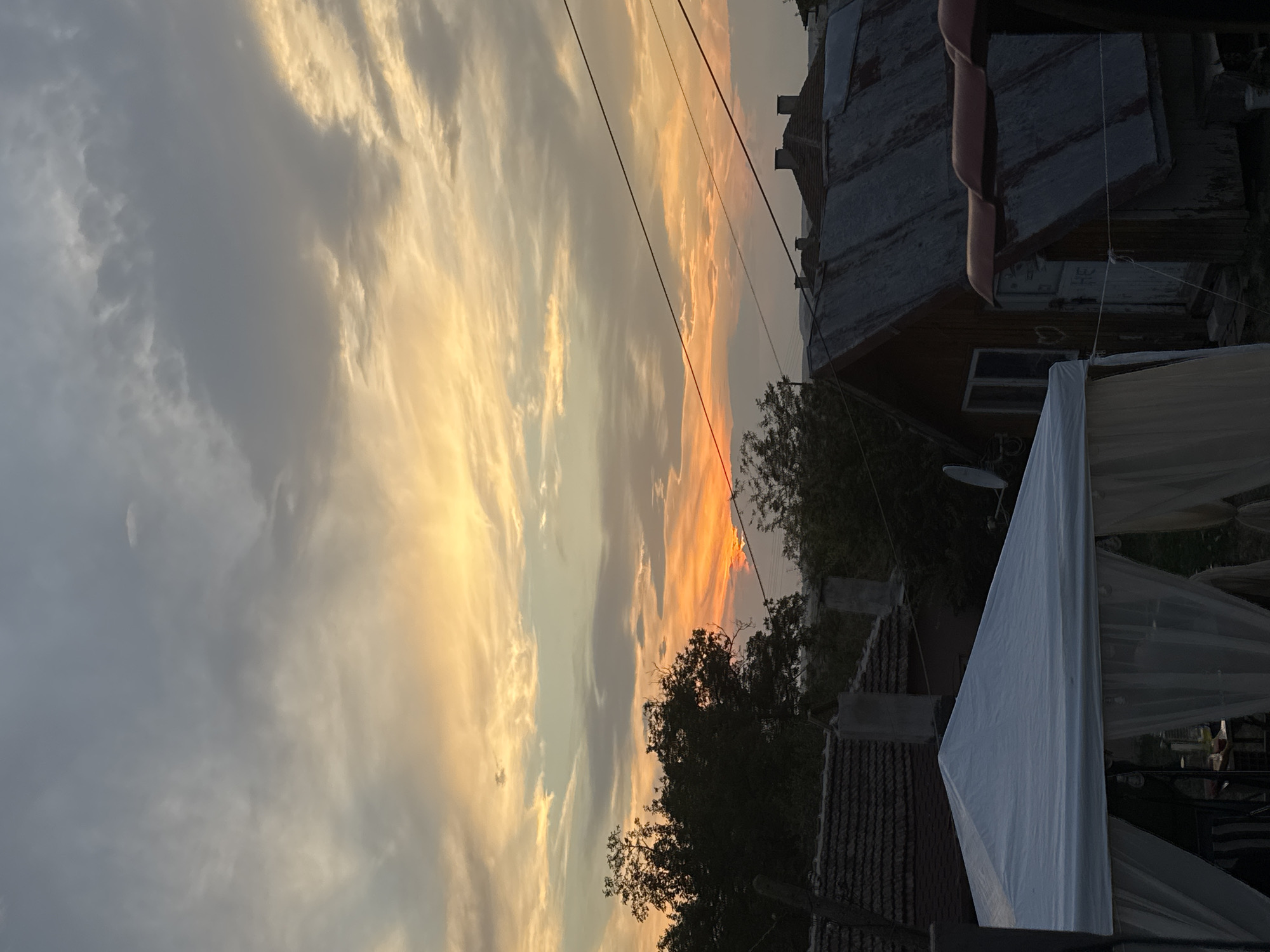 This image captures a sunset over a rural scene in Sredets, Bulgaria. The sky is filled with dramatic clouds, painted in hues of orange, yellow, and gray. In the foreground, a white gazebo with sheer curtains is partially visible on the left, suggesting an outdoor gathering or event. Behind it, several rustic buildings with corrugated metal roofs are scattered across the landscape. Trees with dark foliage are silhouetted against the vibrant sky. Electrical wires stretch across the scene, indicating a rural infrastructure. The overall impression is one of a peaceful evening in a Bulgarian village.