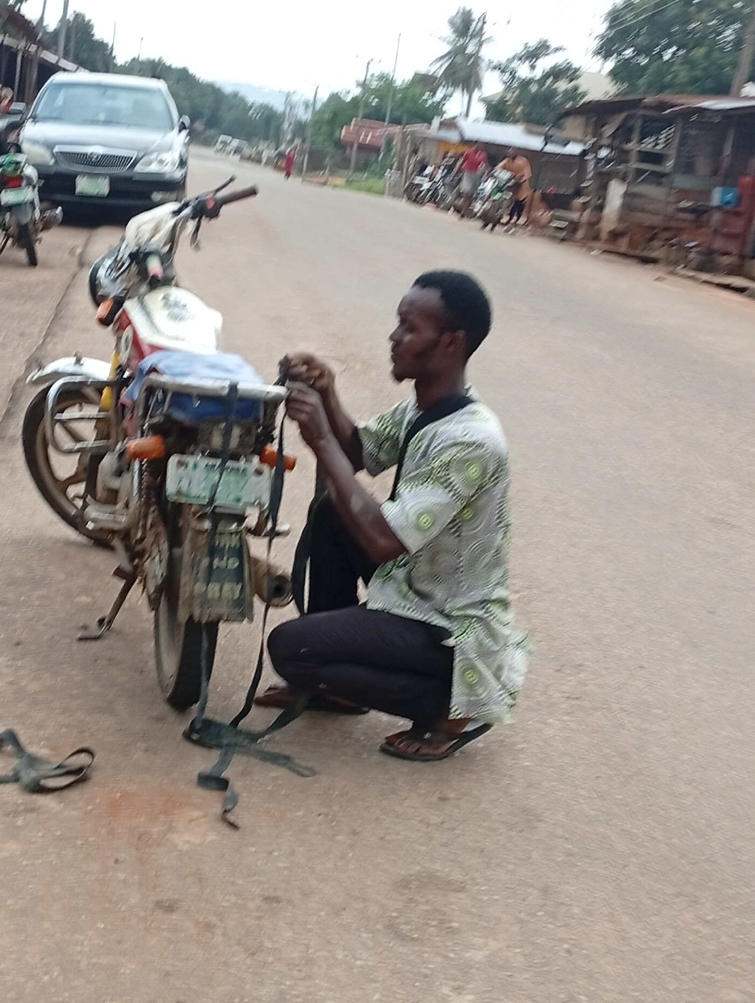 In Nsugpe, Nigeria, a young man is seen squatting on an asphalt road next to a motorcycle. He is wearing a light-colored shirt with a green circular pattern and dark pants. The motorcycle is parked on its kickstand and appears to be partially disassembled or being worked on. A dark strap lies on the ground near the motorcycle. In the background, a silver sedan is parked, and several other motorcycles are visible, along with market stalls and structures lining the road. The sky is overcast.
