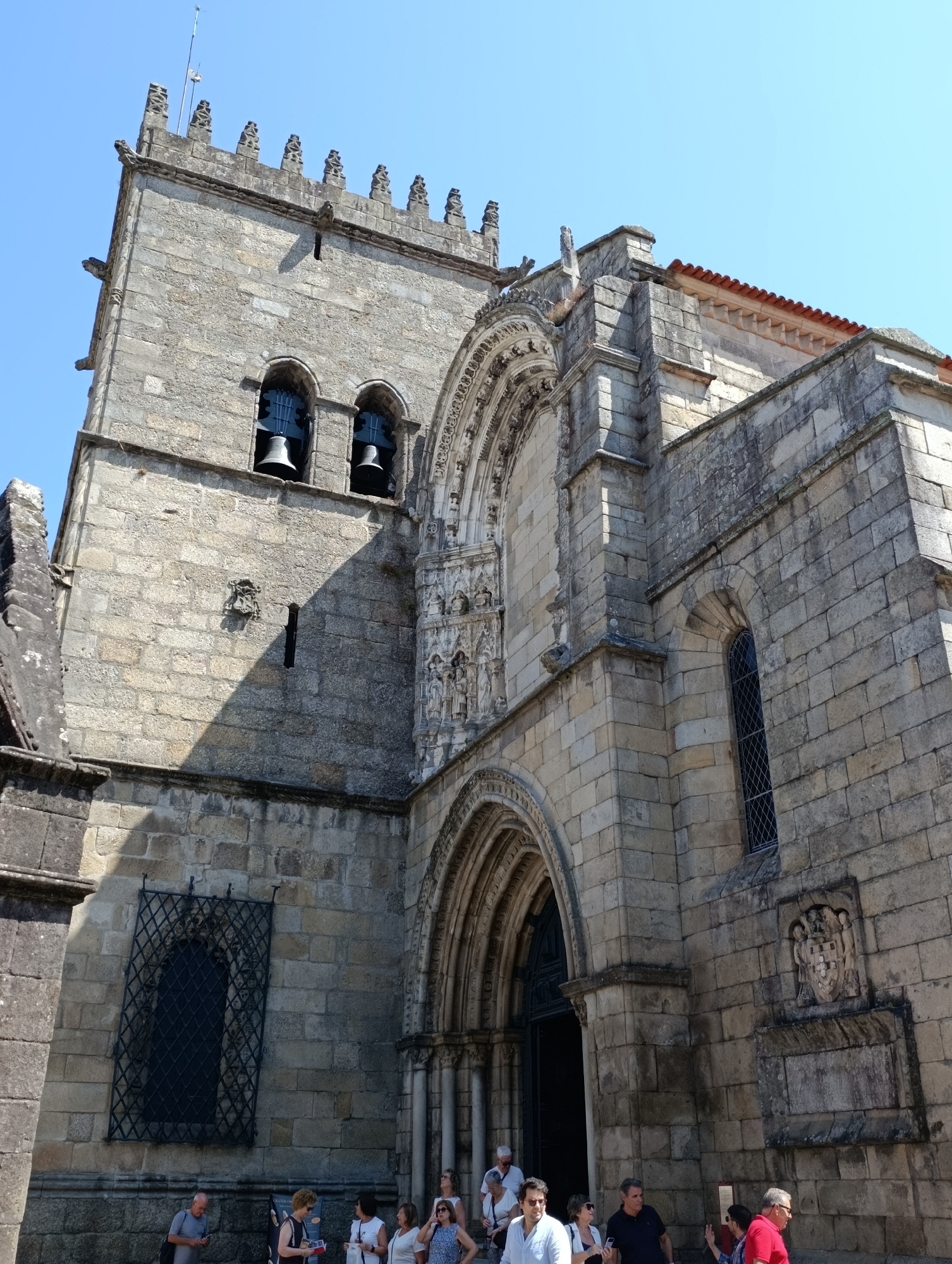 This is a low-angle, eye-level, full shot of the Sé Cathedral of Guimarães, Portugal, on a clear, sunny day. The stone facade of the cathedral is prominent, featuring a large bell tower with two bells visible through arched openings. The tower has crenellated battlements and gargoyles. Adjacent to the tower is a large, ornate stone portal with carved figures and intricate architectural details. A smaller arched window with a dark lattice is visible on the right side of the facade, below a tiled roof. Several people are gathered in front of the cathedral entrance and along the side. Some are looking at their phones, while others are talking or observing the building. The ground in front of the cathedral is paved. The environment is urban, with a clear blue sky above.
