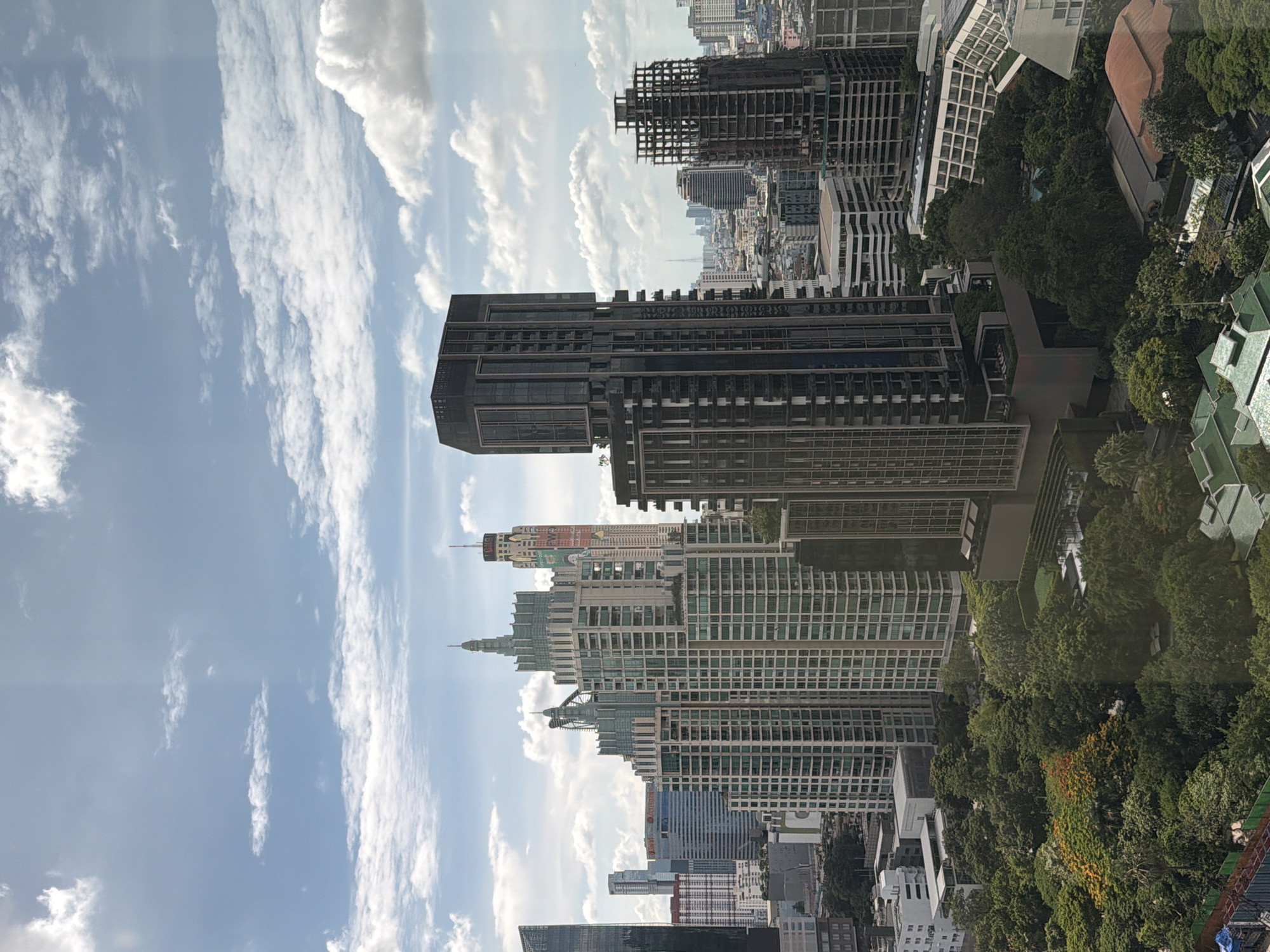 This is a daytime, eye-level, wide shot of a densely populated urban cityscape in Bangkok, Thailand. The foreground is dominated by lush green trees and foliage, with some areas featuring flowering trees with orange and red blossoms. Several buildings are visible within this green space, including some with green tiled roofs and others that appear to be part of a resort or complex.
Behind the trees, the mid-ground and background reveal a dense collection of modern high-rise buildings of varying architectural styles and heights. Notable structures include several prominent skyscrapers with intricate facades, some featuring stepped designs and spires. One particularly striking building is a dark, rectangular skyscraper with many windows and balconies. Another is a building under construction, with its skeletal frame exposed. In the distance, more buildings extend towards the horizon, interspersed with glimpses of infrastructure like antennas and communication towers.
The sky is a clear blue with scattered white clouds, suggesting fair weather. The overall impression is one of a bustling, modern city with pockets of significant green space, likely a park or managed landscape. There are no people or animals visible in the frame.