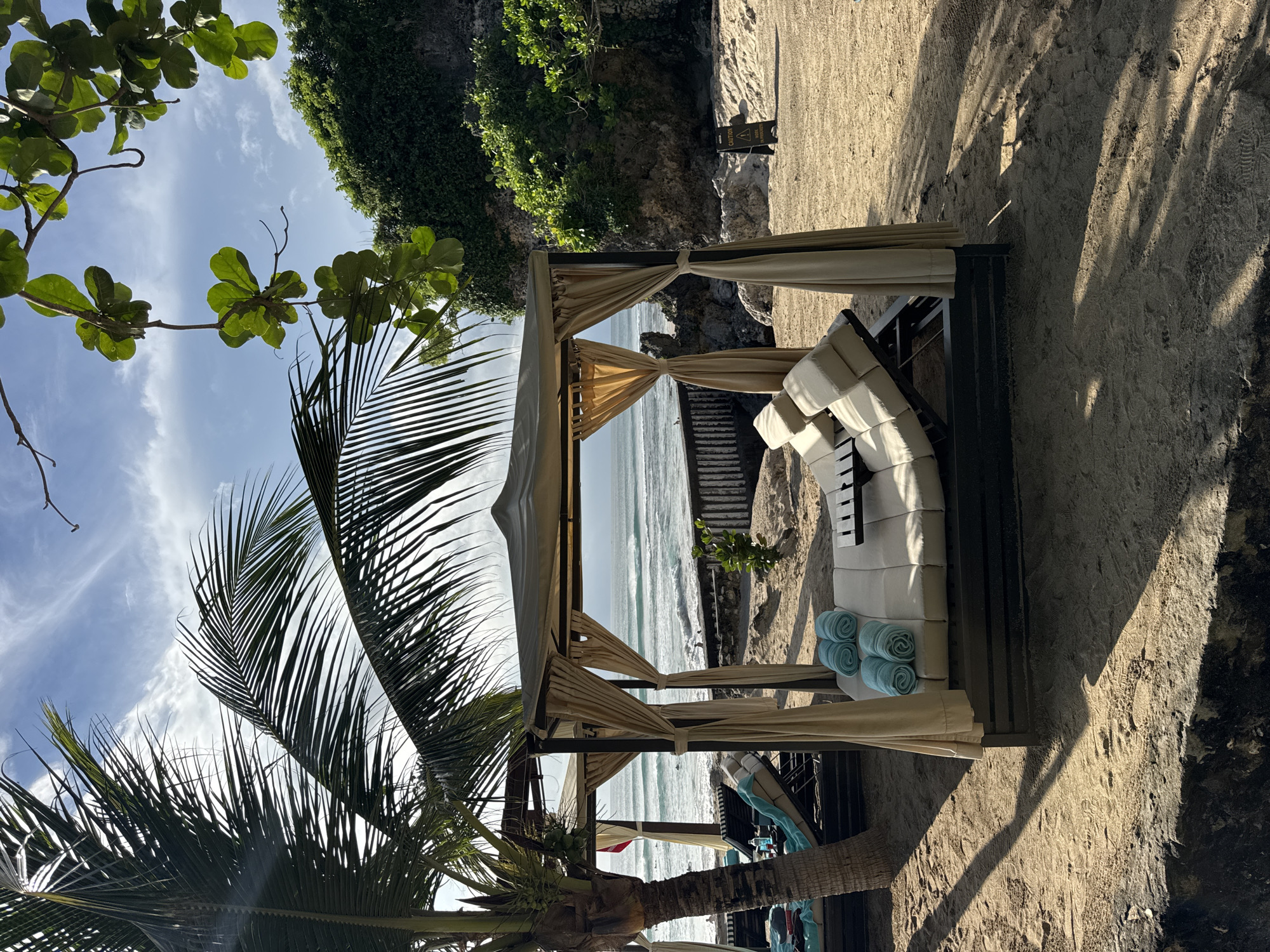 This image, captured in Kuta Selatan, Indonesia, depicts a serene tropical beach scene during daylight hours, likely mid-morning or late afternoon given the shadows. The sky is bright blue with scattered white clouds, framed by lush green palm fronds and other leafy branches in the foreground.
The central subject is a luxurious, open-sided beach cabana, constructed with a dark frame and draped with light beige fabric curtains, tied back to offer an unobstructed view of the ocean. Inside, a comfortable white lounge chair or daybed, adorned with multiple plush cushions, invites relaxation. Several light blue rolled towels are neatly stacked on the right side of the lounge chair, ready for use.
Beyond the cabana, the calm, blue ocean stretches towards the horizon, with gentle waves breaking near the sandy shore. The beach itself is finely textured, bearing subtle patterns and long shadows cast by the cabana and surrounding elements. To the right, a rocky cliff face rises, densely covered in vibrant green foliage. A dark, possibly wooden, staircase or pathway is partially visible, appearing to descend towards the water from the cliff area. Near the cliff, a small black and yellow triangular warning sign is visible, displaying the vertically oriented text "CAUTION" and "SLIPPERY."
The scene exudes an atmosphere of peaceful relaxation and tropical tranquility. There are no people visible, emphasizing the untouched beauty and exclusive feel of the location.