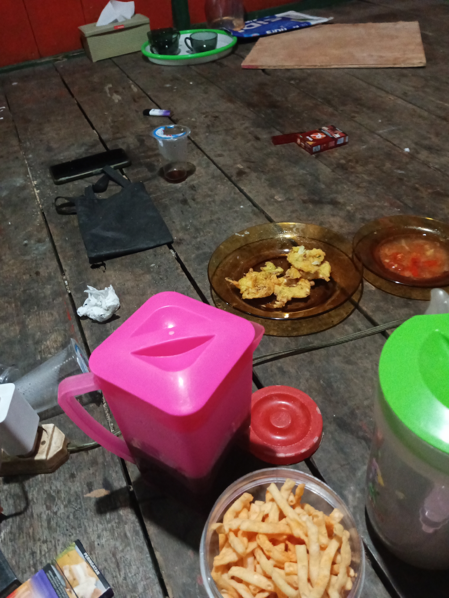 The image depicts a cluttered wooden table with various objects, suggesting a casual setting. In the foreground, a pink pitcher with a lid is prominent, next to a red circular object. To the right, a clear container filled with small, golden fried snacks is visible, with a large green-lidded container partially visible behind it. Further back, two small amber-colored plates hold fried food and a red sauce. To the left, a black tote bag and a smartphone lie on the table. A crumpled piece of white paper is near the pitcher. In the background, a tissue box sits on a red surface, accompanied by two dark cups on a tray. A cigarette pack is also present on the table. The overall impression is one of a relaxed, informal gathering. The mention of Tumampua, Indonesia is not supported by visual evidence in the image.