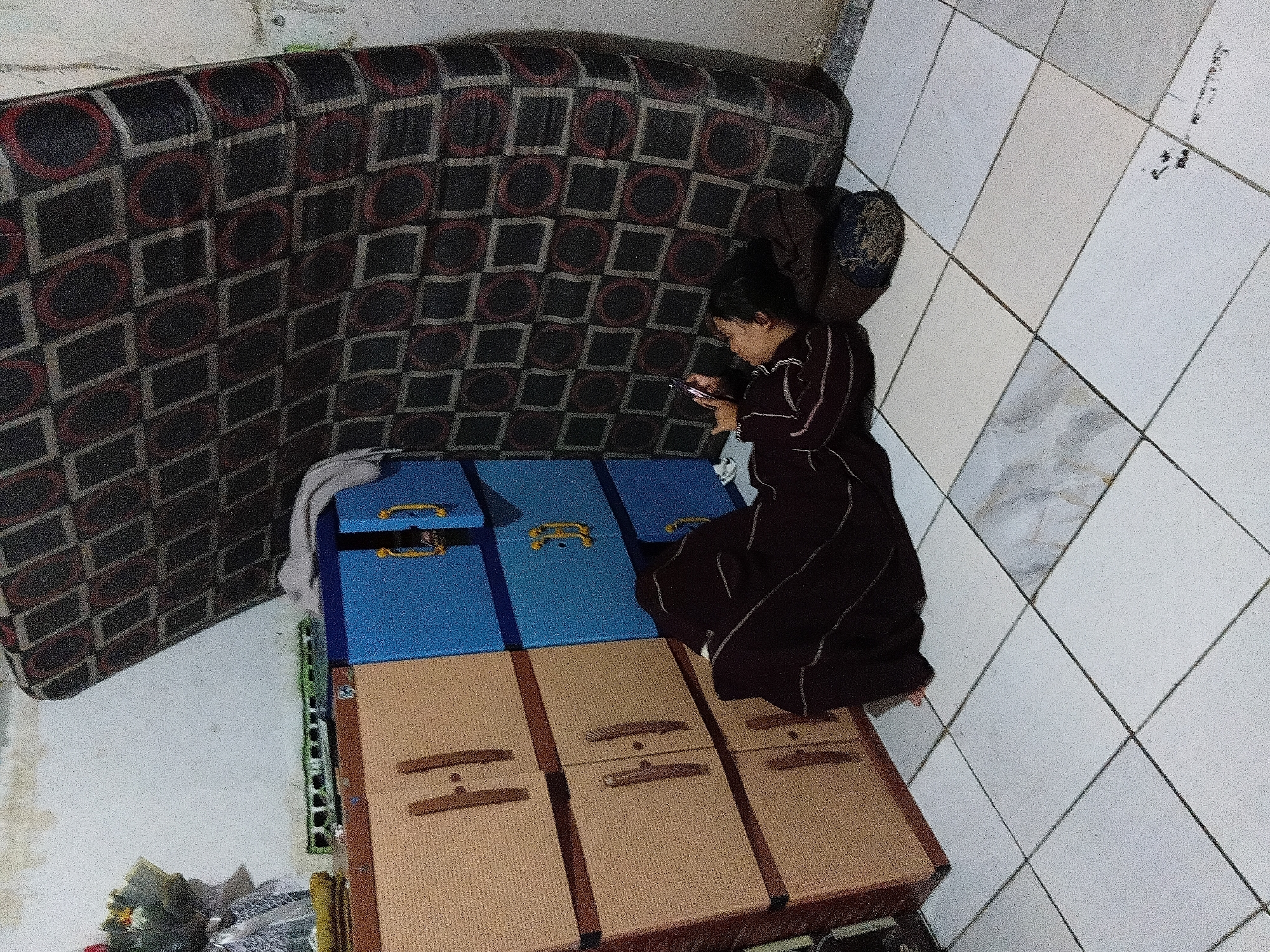 The image depicts an indoor scene, possibly a residential room, in Bekasi, Indonesia. In the foreground, a person is lying on their back on the tiled floor, using a mobile phone. They are wearing a dark, striped garment. Behind the person, a large mattress leans against a wall. To the left of the person and mattress is a multi-drawer cabinet, colored in beige and blue. A decorative bouquet sits on top of the beige drawers, and a toy train is placed nearby. The environment appears somewhat cluttered, with visible wall texture and floor tiling.