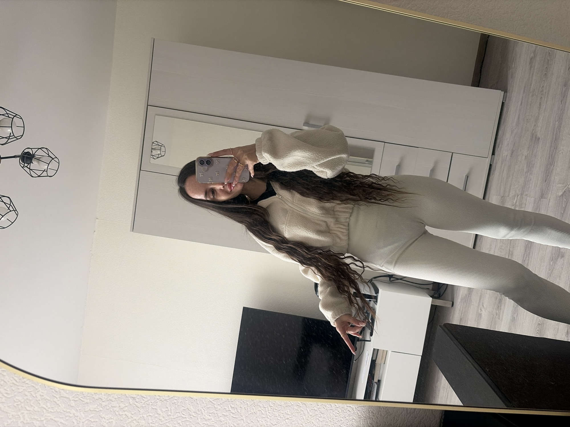 The image shows a woman taking a selfie in a mirror. She is wearing a fuzzy off-white jacket and matching ribbed leggings. The background includes a white wardrobe, a television, and a black coffee table. The location does not appear to be in Saint Petersburg, Russia.