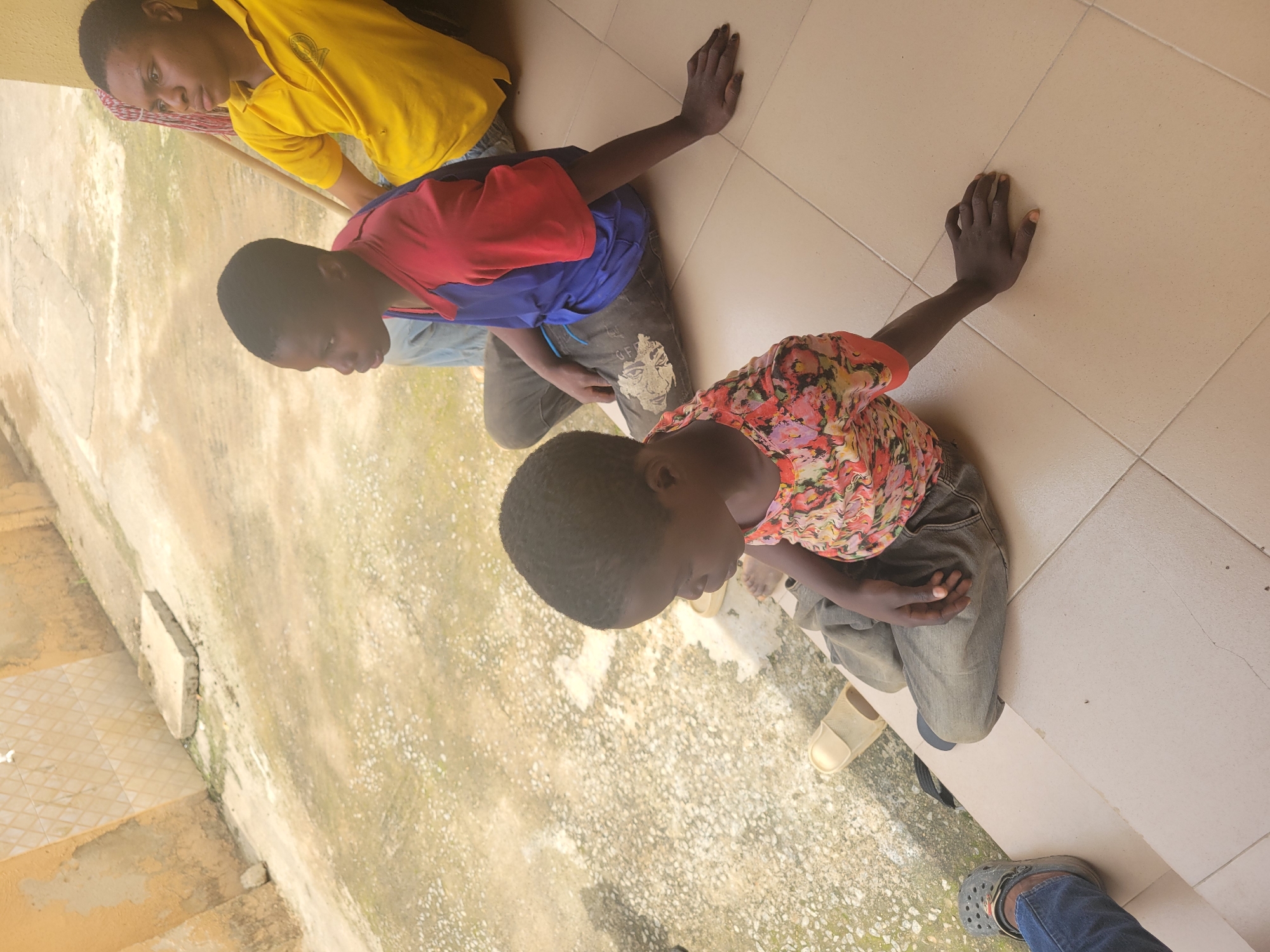 Three young people are sitting on a tiled porch outside a building with a textured, ochre-colored wall. The person in the foreground, on the left, is wearing a colorful floral-patterned t-shirt and dark pants. Their left arm is resting on the tiled floor, palm down. To their right, another young person is wearing a blue and red t-shirt and dark pants. Their right arm is also resting on the tiled floor. Further to the right, a third young person is wearing a bright yellow polo shirt. They are looking to the right. The ground outside the porch is rough and uneven, with some patches of greenery. A mop handle is visible behind the person in the yellow shirt. The scene appears to be outdoors, in what could be a residential or community area.