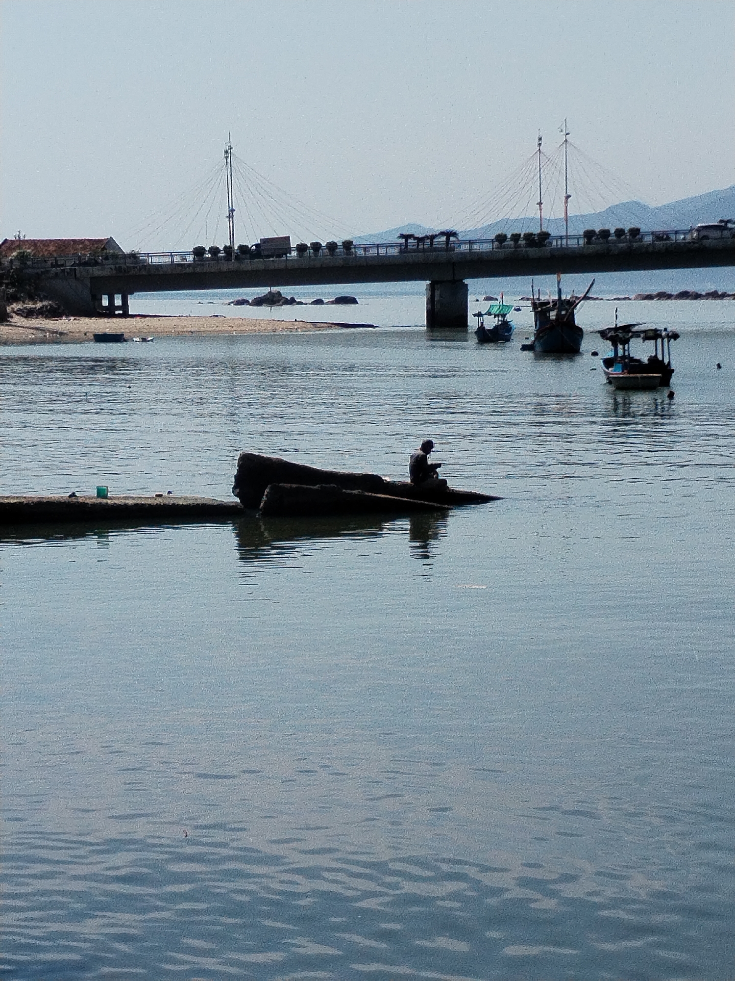 Captured from a distance, the image showcases a serene waterfront scene in Bac Nha Trang Ward, Vietnam. Predominantly composed of calm, blue water, the foreground features a partial concrete structure extending from the left edge, adorned with a green container. A dark-colored, weathered tree trunk sits adjacent, serving as a perch for a solitary individual in dark attire, seated and seemingly engaged in an activity.
In the middle ground, several small, traditional Vietnamese boats are anchored, gently bobbing on the water. A bridge, adorned with planted greenery along its span, stretches across the background. Vehicles traverse the bridge, and distant hills provide a soft, natural backdrop. The sky is pale and clear, suggesting a bright, cloudless day. The overall atmosphere is tranquil and showcases a typical coastal scene in Vietnam.