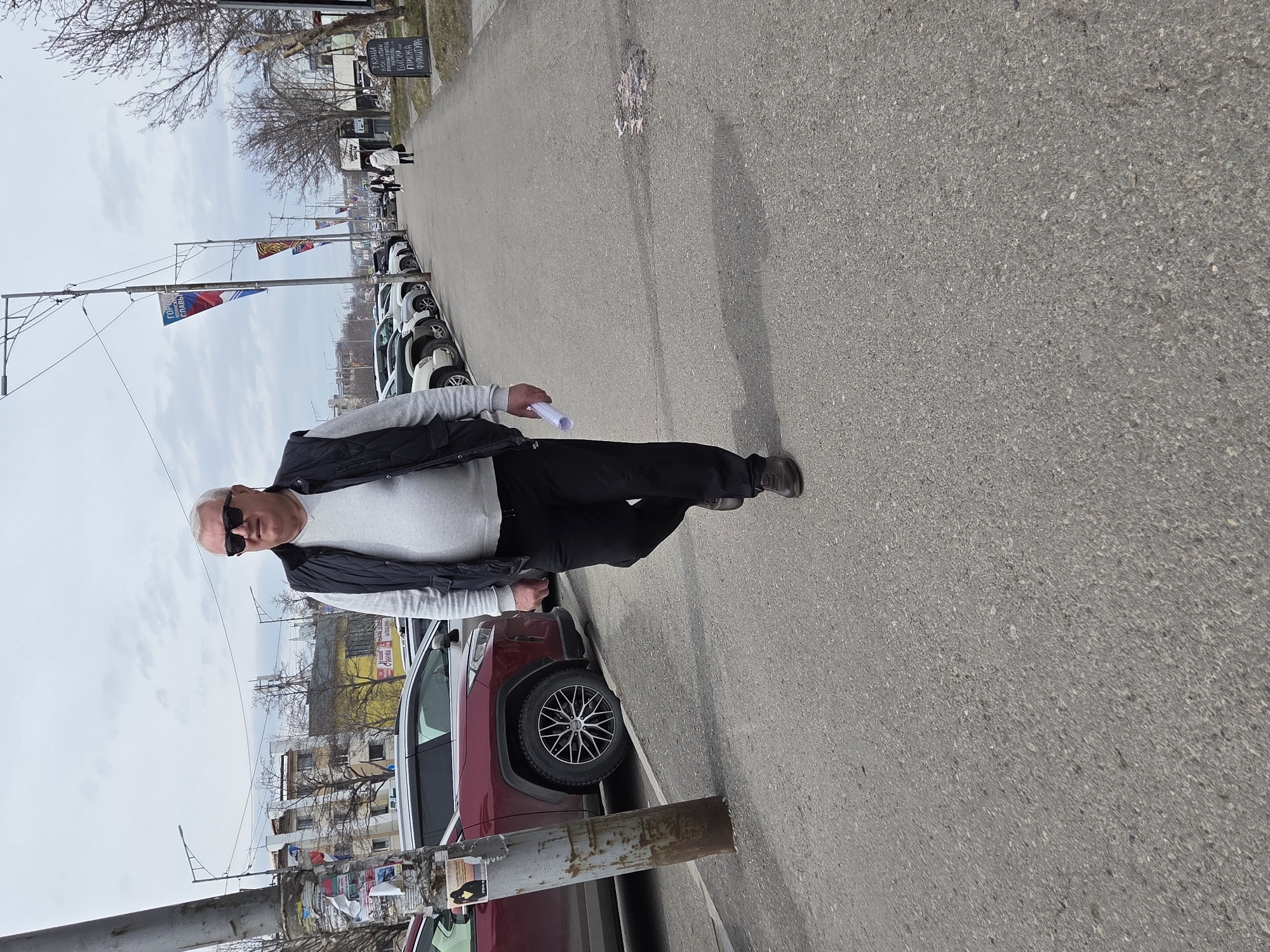 A man with white hair, sunglasses, a gray sweater, a black vest, and black pants is standing on a city street. He is holding a rolled-up paper in his left hand. A row of cars is parked on the left side of the street. There are flags on poles and buildings in the background. The street is paved with asphalt.