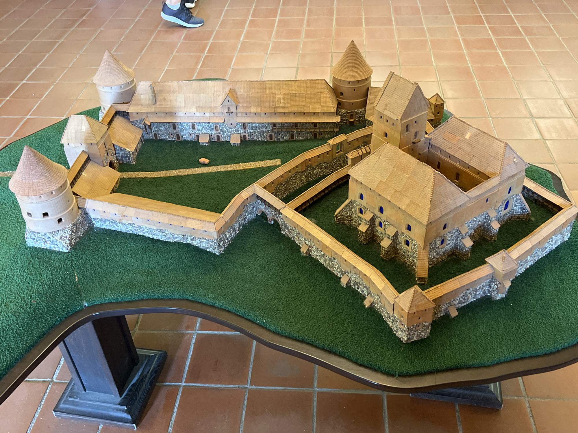 A detailed scale model of Trakai Island Castle, located in Trakai, Lithuania, is displayed on a dark-brown, curved table. The model features a sprawling complex of wooden and stone structures, including several towers with conical roofs, a large central courtyard, and extensive defensive walls. The model is situated on a green, grassy base, designed to represent the surrounding landscape. The table is placed on a floor tiled with reddish-brown squares. In the upper left portion of the image, the lower legs and feet of a person wearing athletic shoes are visible, indicating their presence near the display. The lighting appears to be natural, coming from above.