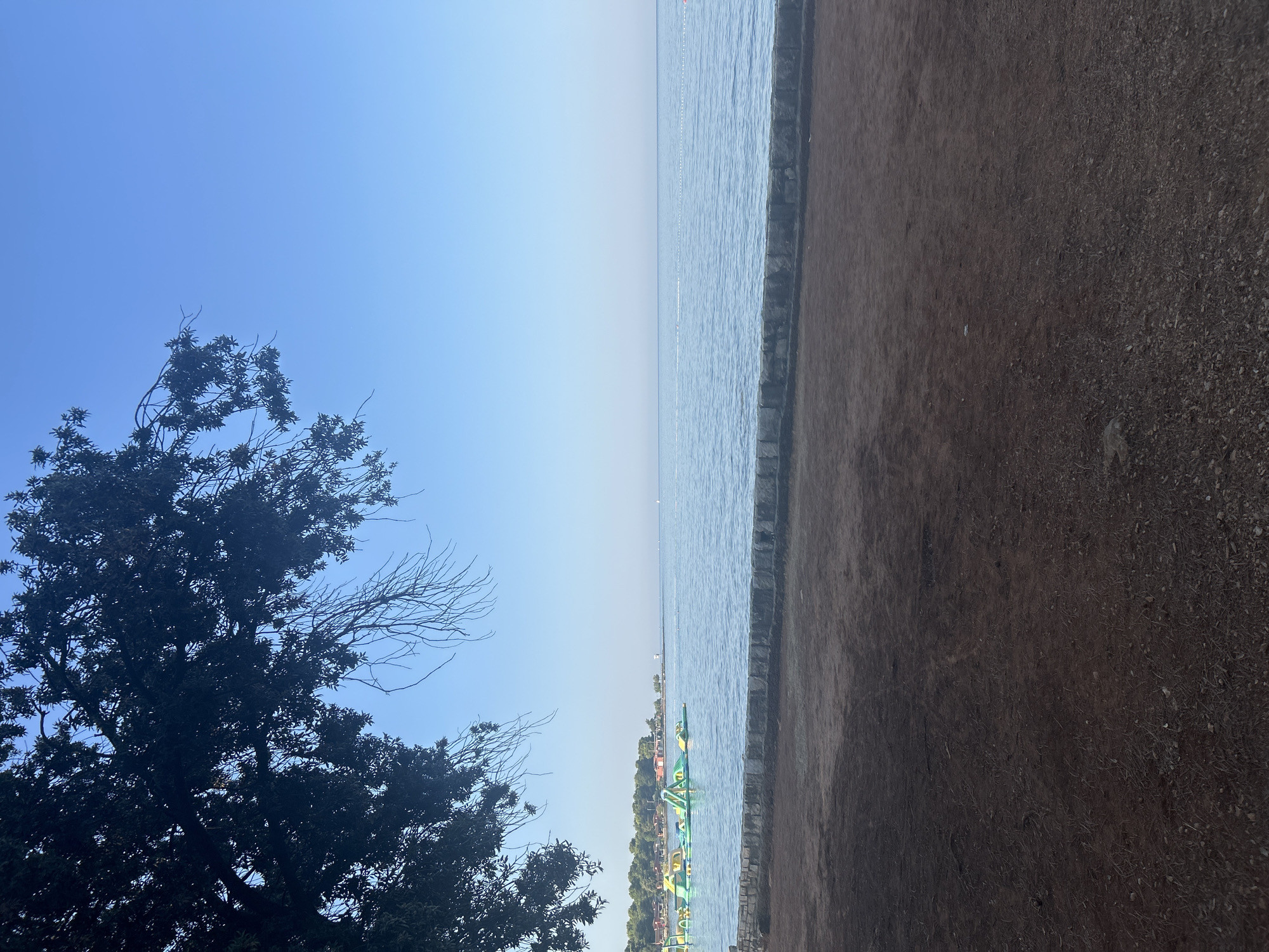 A serene coastal scene unfolds under a clear, pale blue sky. The foreground is dominated by a dark, earthy expanse, possibly a beach or a dirt path, with sparse vegetation and small stones scattered across its surface. A low, textured stone wall separates this area from the tranquil, calm sea. The water itself is a soft blue, reflecting the sky, and a thin white line, possibly buoys or a marker, stretches across it.
In the middle distance, a vibrant inflatable water park with yellow and green structures rests on the surface of the sea. Beyond the water park, a tree-lined coast is visible, with hints of buildings and structures nestled amongst the greenery. The foliage of a large, dark tree casts a shadow in the upper left quadrant of the image, its branches reaching towards the sky. The overall impression is one of peacefulness and summer recreation. The location is identified as being in Monterol, Croatia.