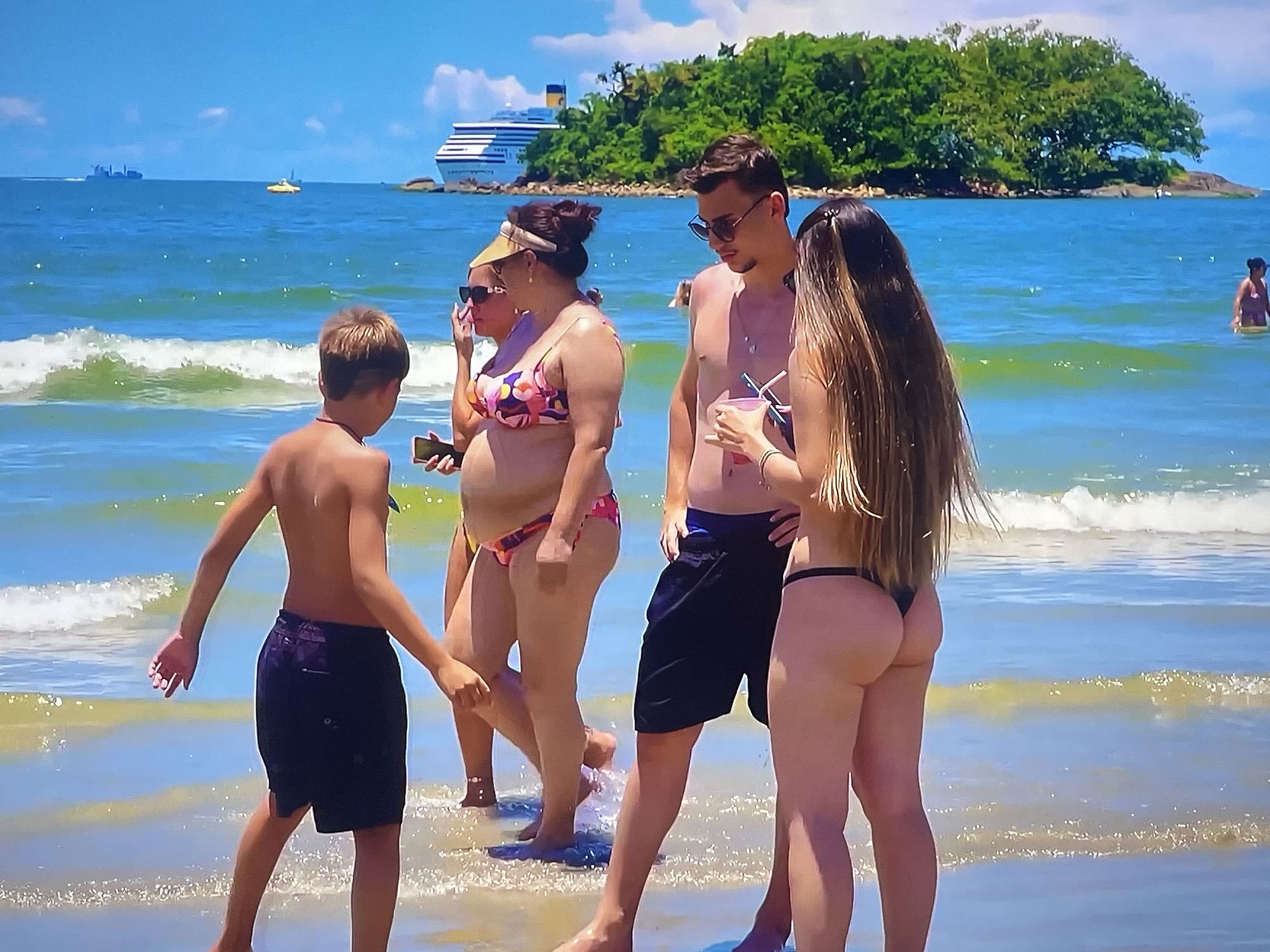 In a sun-drenched outdoor scene, a group of people enjoys a day at the beach. The water is calm and clear, with gentle waves lapping at the shore. The sky is a brilliant blue, dotted with a few fluffy white clouds. In the distance, a large cruise ship is visible, adding to the vacation atmosphere. The group consists of at least four individuals, including two men, a woman, and a boy. They are all dressed in swimwear, suggesting a casual day of relaxation and fun. The woman is wearing a floral bikini, while the others are in swim trunks and bikinis.
The overall mood of the scene is one of leisure and enjoyment. The bright sunlight and the presence of the ocean evoke a sense of freedom and escape. The people appear to be interacting with each other, possibly chatting or enjoying each other's company. The boy, in particular, seems to be exploring the water's edge, his hands extended as if reaching for something.
The backdrop of the scene is a lush green island, hinting at a tropical or subtropical location. The presence of a large cruise ship further suggests a popular tourist destination. The time of day appears to be midday or early afternoon, given the intensity of the sunlight. The weather is clearly warm and pleasant, perfect for a beach outing. There is no visible text in the image. The specific location is Shakhovskaya, Russia, which adds a unique geographical context to this otherwise universal beach scene.