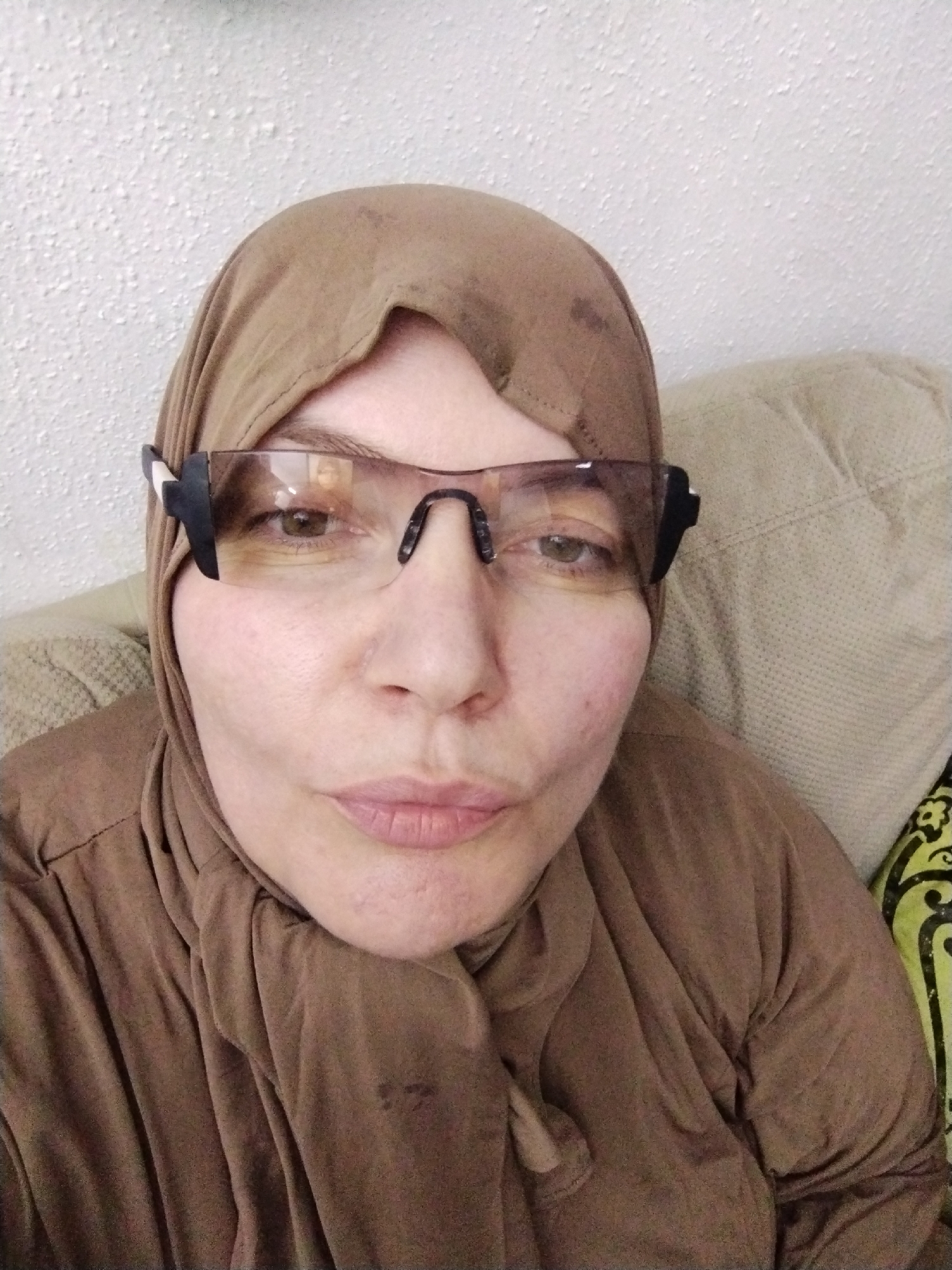 A woman wearing a brown hijab and brown-tinted glasses is looking directly at the camera. The background is a white wall and a beige couch. The woman has fair skin and appears to be indoors. There is no indication of the city of Al Mas'oudi, United Arab Emirates, or any other specific location.