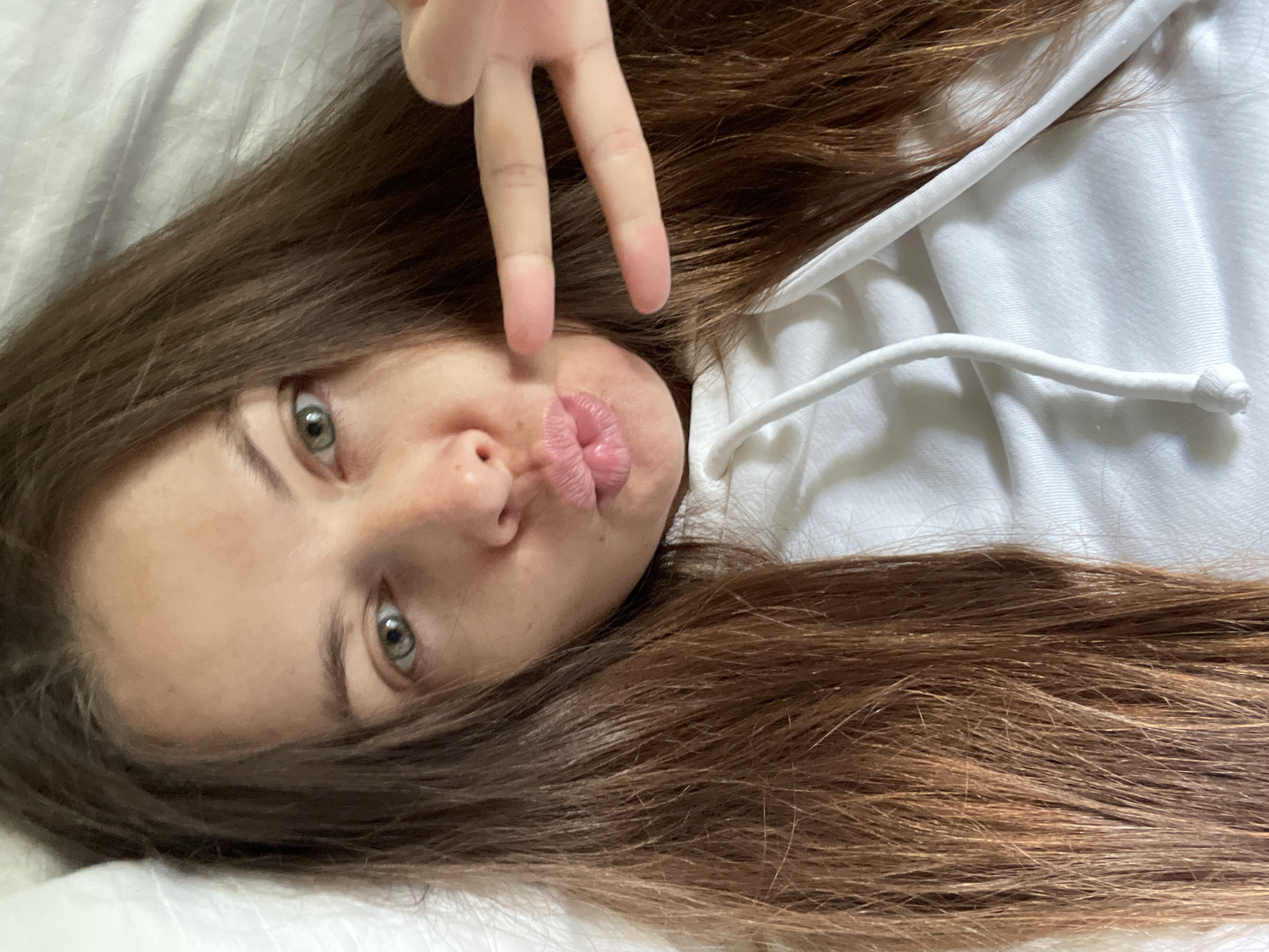 The image shows a close-up, overhead, selfie-style shot of a woman lying in bed. She has long, brown hair spread around her face and shoulders. Her eyes are open and looking towards the camera, and her lips are pursed in a kiss. Her right hand is raised, with two fingers extended in a peace sign. She is wearing a white hooded sweatshirt. The background appears to be a white bedsheet and pillow. There is no indication of the city of Korotycha, Russia, or any other specific location.