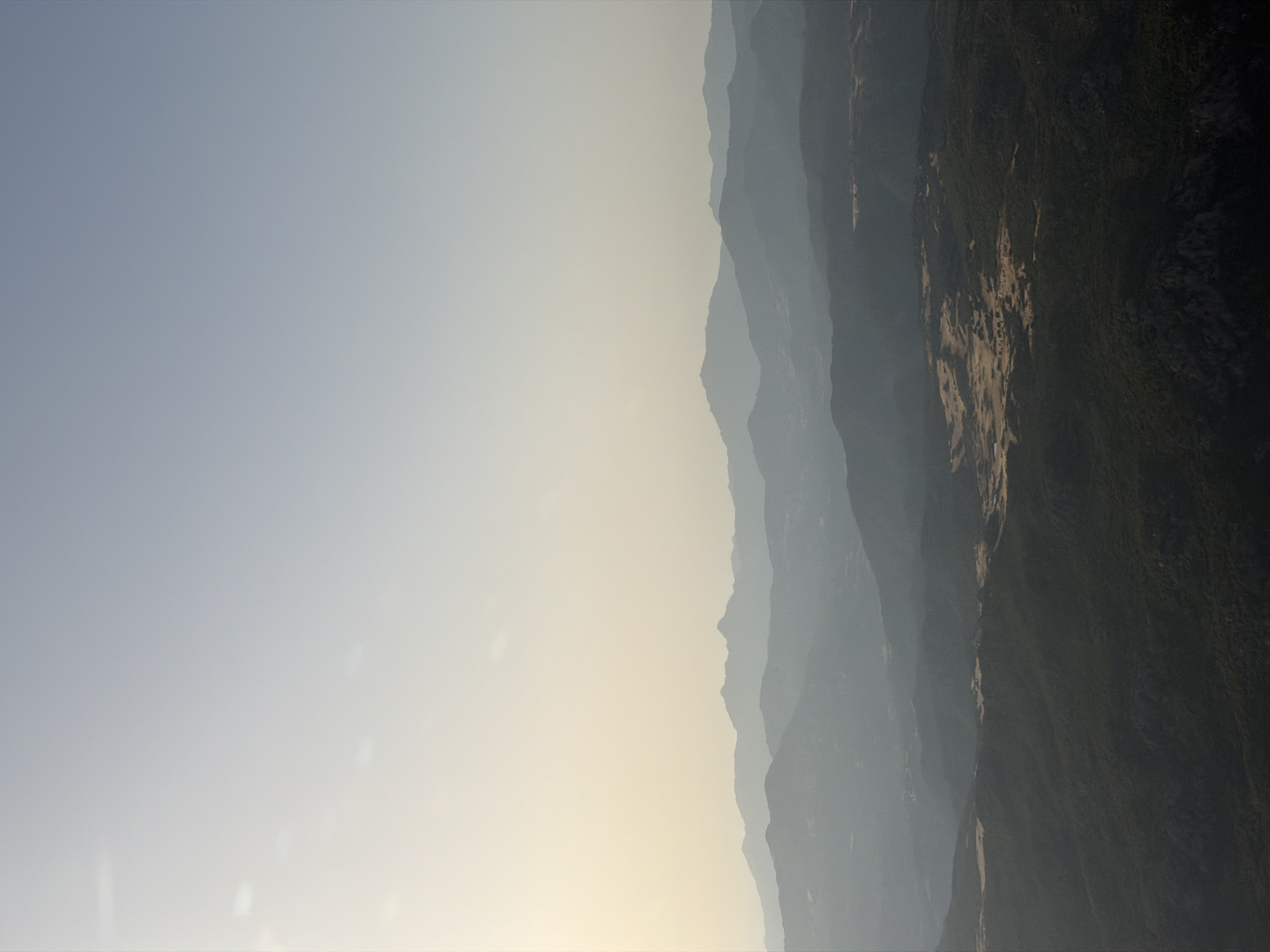 This is an aerial photograph of a mountainous landscape near Monte/Monti, Italy. The image features multiple layers of hazy, silhouetted mountains fading into the distance. The sky is a pale, clear blue, transitioning to a soft yellow near the horizon, suggesting either early morning or late afternoon light. The foreground is a dark, densely vegetated mountainside, with patches of lighter, dry vegetation or clearings visible on a plateau further into the scene. There are no people or man-made structures immediately discernible in the image, focusing solely on the natural topography.
