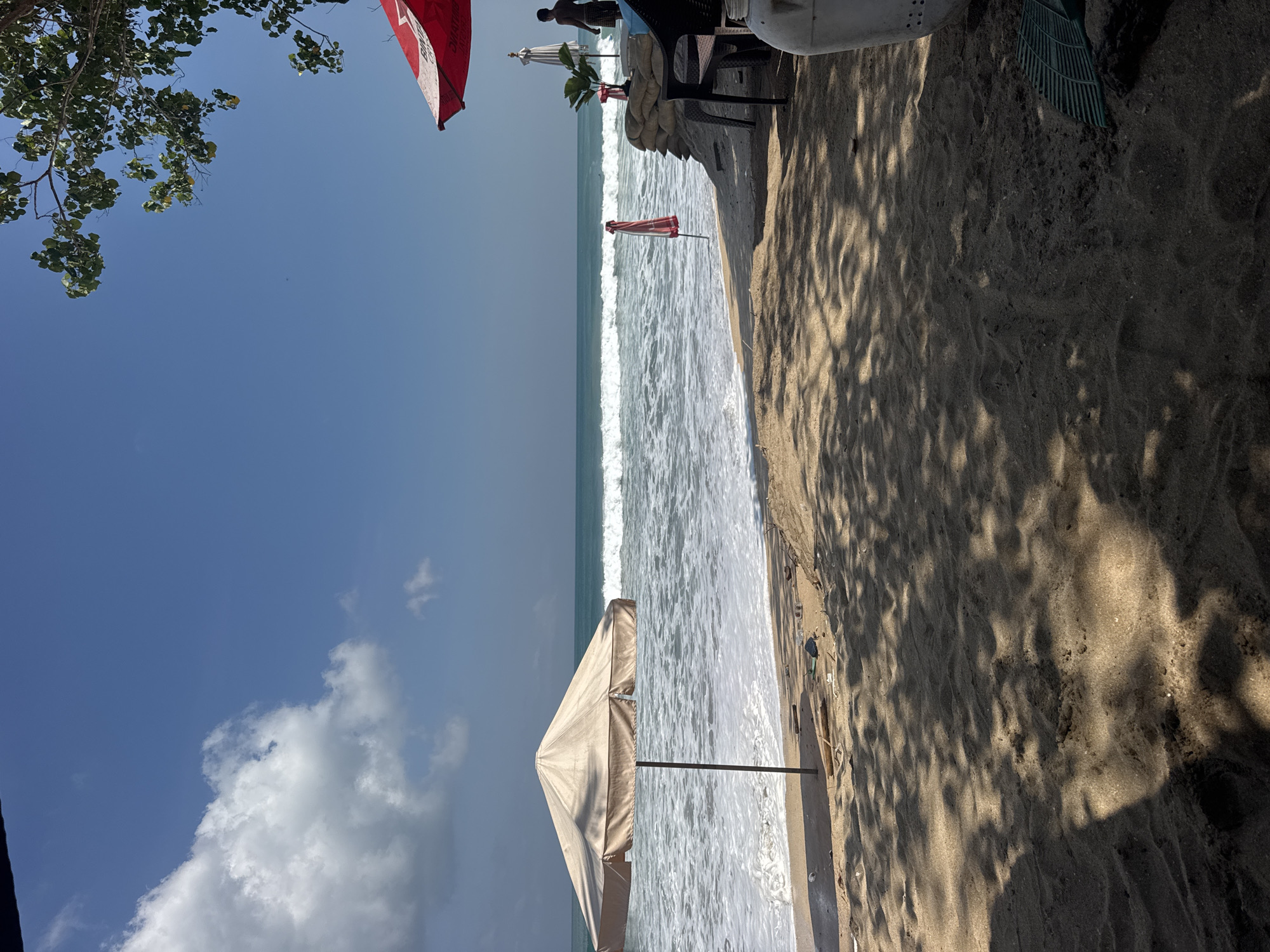 The image depicts a beach scene in Kuta, Indonesia, on a clear day. In the foreground, dark sand is partially shaded by tree branches, creating dappled patterns. A green rake lies on the sand to the right. In the midground, a beige beach umbrella stands on the sand near the water's edge. The ocean features moderate waves with white foam crashing onto the shore. Further to the right, a large sandbag barrier is visible, with a person standing behind it, wearing dark shorts and a light-colored shirt. Above this barrier, a red and white umbrella is partially visible. A small green plant sits on top of the sandbags. In the background, the turquoise ocean extends to the horizon under a bright blue sky with scattered white clouds.