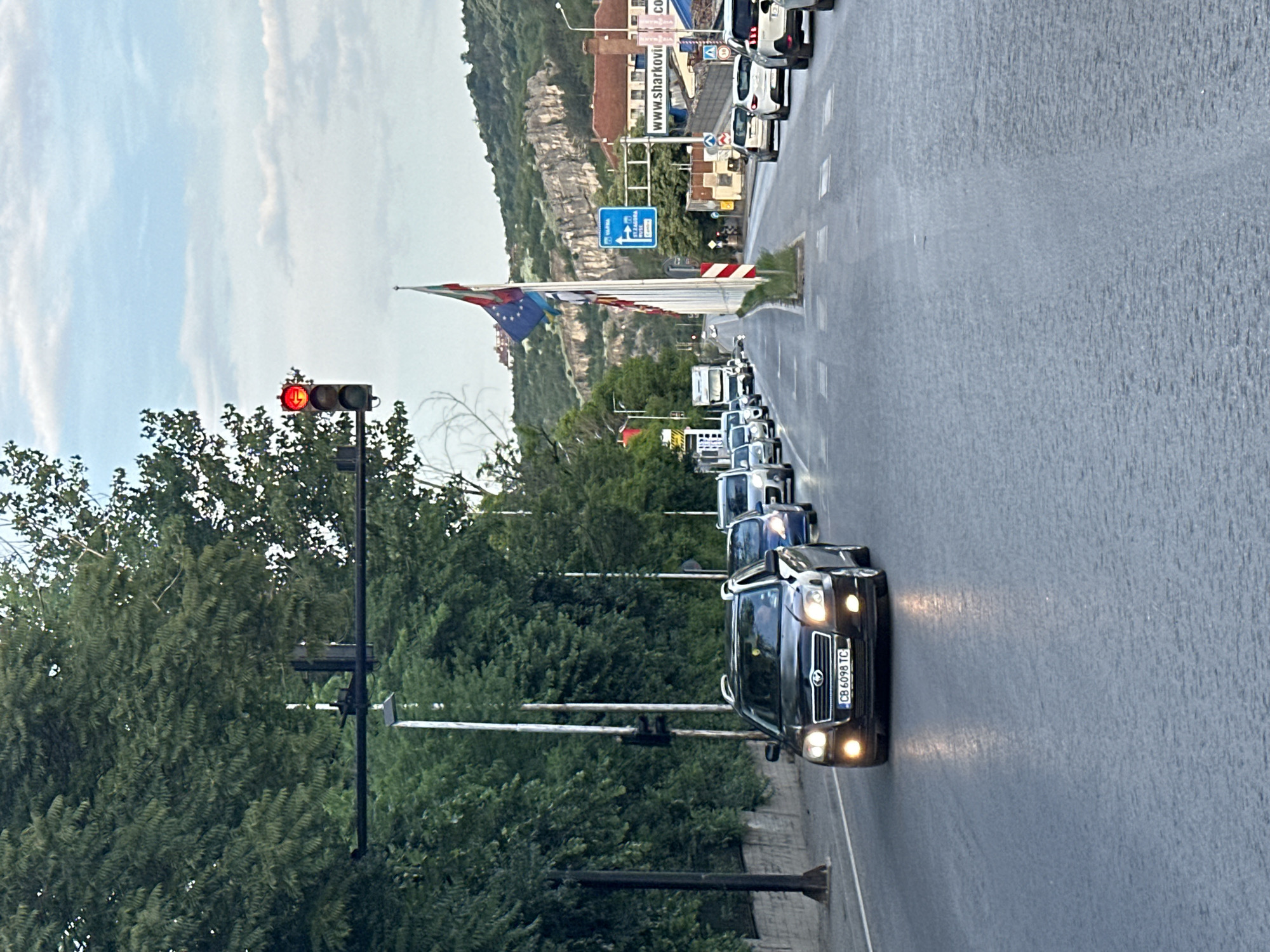 The image captures a road scene, likely in Veliko Tarnovo, Bulgaria, given the provided context. The perspective is from inside a vehicle, looking forward along a moderately busy road.
In the foreground, a dark-colored SUV (possibly a Toyota) is visible, its headlights illuminated, indicating it's either in motion, preparing to move, or the lights are on. Its Bulgarian license plate, "CB 6098 TC", is clearly legible, with "CB" signifying Sofia region registration. The road surface appears wet, suggesting recent rain or damp conditions.
Further down the road, a line of cars is visible, indicating a traffic queue. A red traffic light is prominently displayed on the left, stopping the flow of vehicles. Lush green trees line the sides of the road.
In the background, a distinctive rocky hill or mountain formation rises, and various buildings are scattered along its base. A row of flagpoles stands tall on the right side of the road, displaying multiple national flags, most notably the European Union flag and the Bulgarian flag (white, green, red).
Several road signs are also visible. One blue directional sign contains text in Cyrillic script, consistent with the Bulgarian location. Another sign on a building reads "www.sharkovi.com", with "CO" partially visible beneath it, possibly part of a business name. The sky above is light, with scattered clouds. The overall impression is of a typical daytime urban or semi-urban environment experiencing traffic congestion.