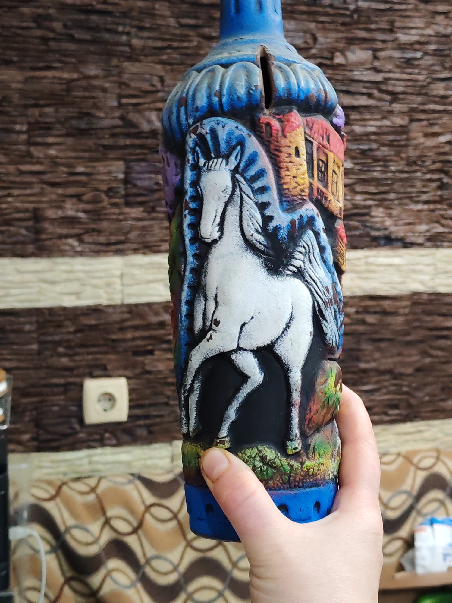 A person is holding a blue, cylindrical, carved ceramic piggy bank. The piggy bank depicts a white horse with wings in the center, a multi-story building to the right, and vegetation at the bottom. The piggy bank has a slot at the top. The person is holding the piggy bank with their right hand. The background includes a brown and white striped wall, an electrical outlet, a couch with a wavy brown and tan pattern, and some items on a table to the left. The image appears to be taken indoors. The location is Unknown, Unknown.