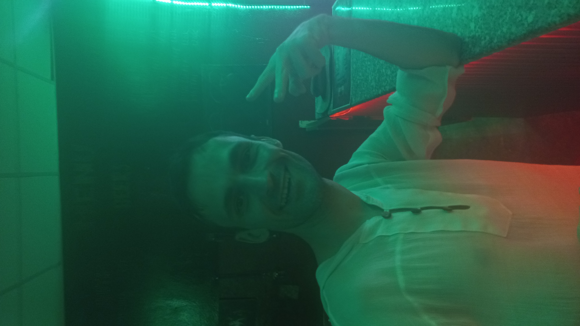 A man is smiling and giving the peace sign in an indoor setting illuminated with green and red light. The location is unclear, but it is in Mũi Né, Vietnam. The man is wearing a light-colored shirt. There is a counter or shelf visible behind him. A dark object, possibly a speaker or electrical panel, is visible on the left.