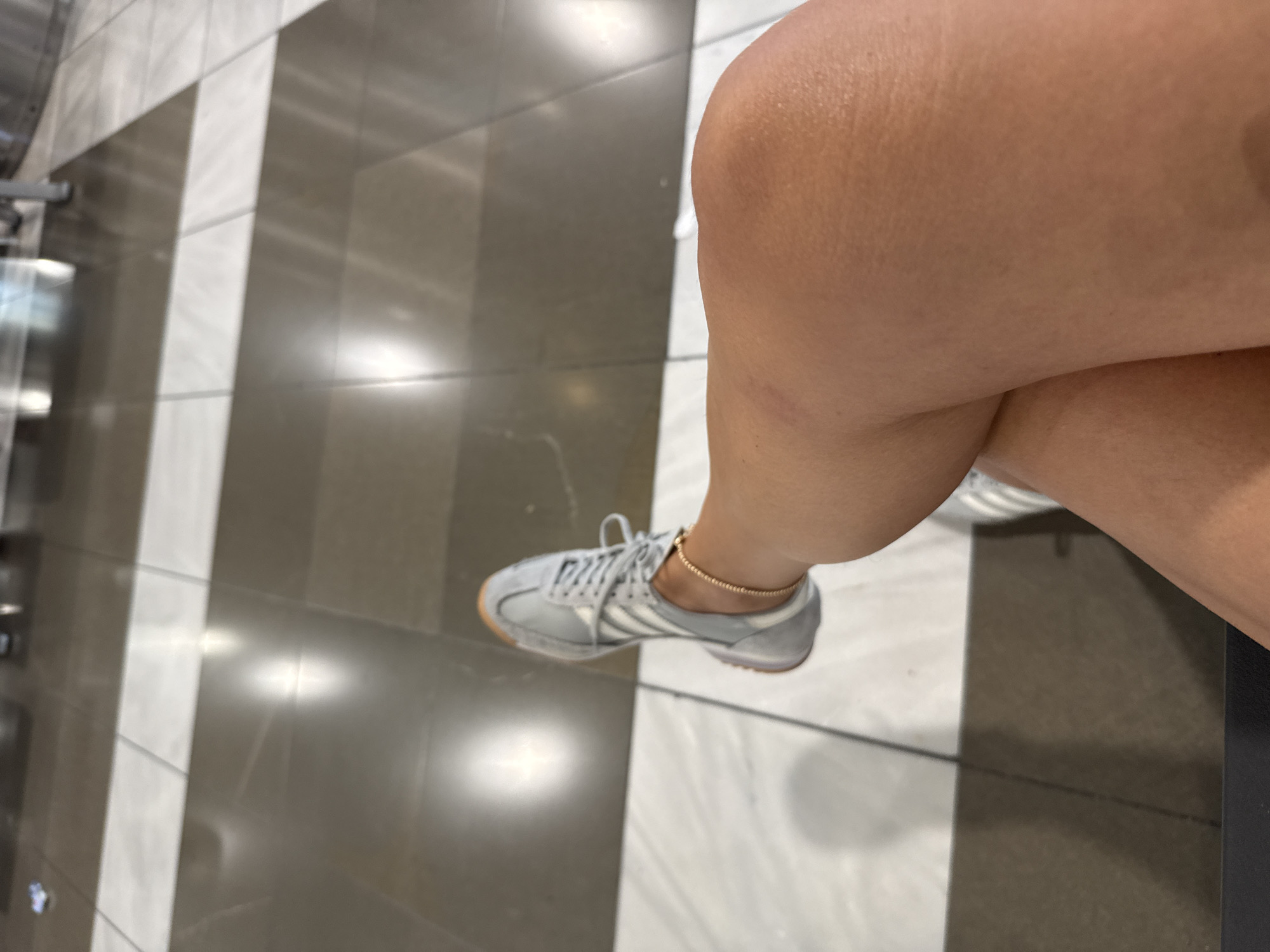 An overhead perspective shows a person's legs and feet from the knees down, seated on a dark bench. The person is wearing grey Adidas sneakers with white stripes and a thin gold ankle bracelet. The floor is tiled in a checkered pattern of light and dark grey squares. The scene is indoors, likely in a public space such as an airport or train station, given the polished floors and lighting. The specific location in the city of Thermi, Greece cannot be determined from the image.