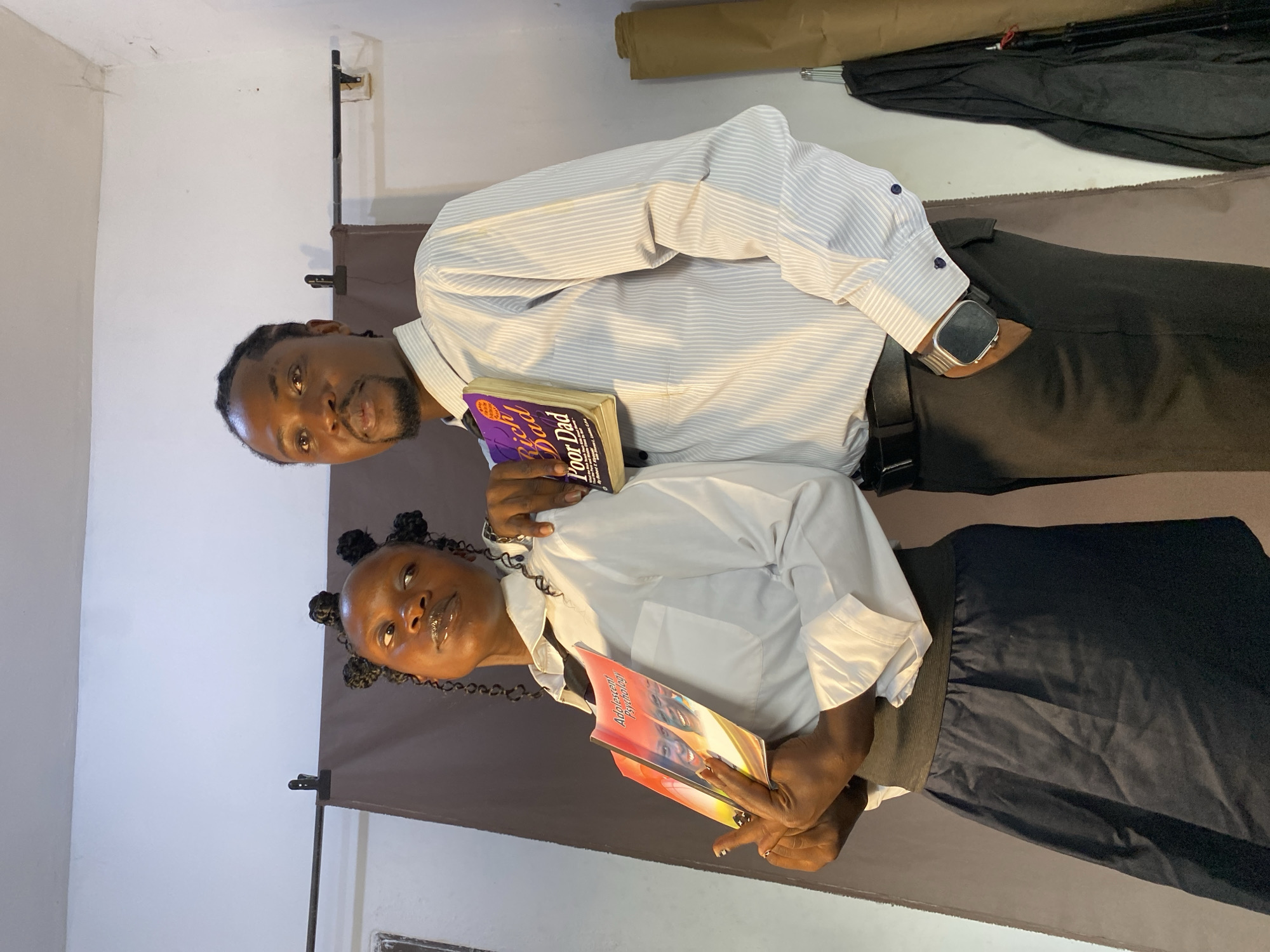 The image, taken in Bukuru, Nigeria, depicts two individuals posing indoors, presented vertically as if rotated 90 degrees clockwise. They appear to be in a makeshift photo studio or a room with a backdrop.
The man, on the left, has dark skin, braided hair, and a neatly trimmed beard. He is wearing a light-colored, thinly striped collared shirt with the sleeves slightly rolled, dark trousers, and a dark-strapped smartwatch on his left wrist. He looks directly at the camera with a neutral expression, holding a copy of the book "Rich Dad Poor Dad" against his chest with his left hand. The purple cover and title are clearly visible.
The woman, on the right, also has dark skin and braided hair styled into several small buns. She wears a white collared shirt and a dark skirt or trousers. She holds a book titled "Adolescent Psychology" in her hands, its colorful cover facing the camera, with a slight smile on her face as she looks forward.
The background consists of a plain white wall on the left and a dark brown fabric backdrop on the right, which is stretched across the frame. Behind the subjects, a rolled-up tan material, black fabric, and metal stands are visible, consistent with a photography setup. The subjects appear calm and composed.