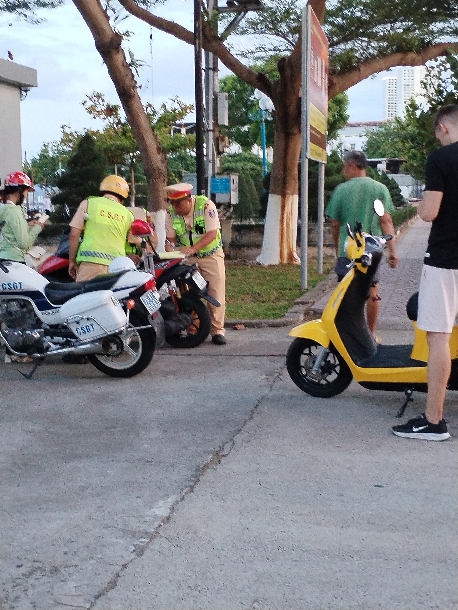 The image depicts a street scene in Nha Trang Ward, Vietnam. Two police officers in khaki uniforms and green reflective vests are interacting with individuals near two parked police motorcycles and a yellow scooter. One officer is writing on a notepad, while the other individuals are observing. One person is wearing a light green jacket and red helmet and holding a cell phone. Another person is wearing a black shirt and white shorts. The environment includes trees, buildings, and a traffic sign, suggesting an urban setting.