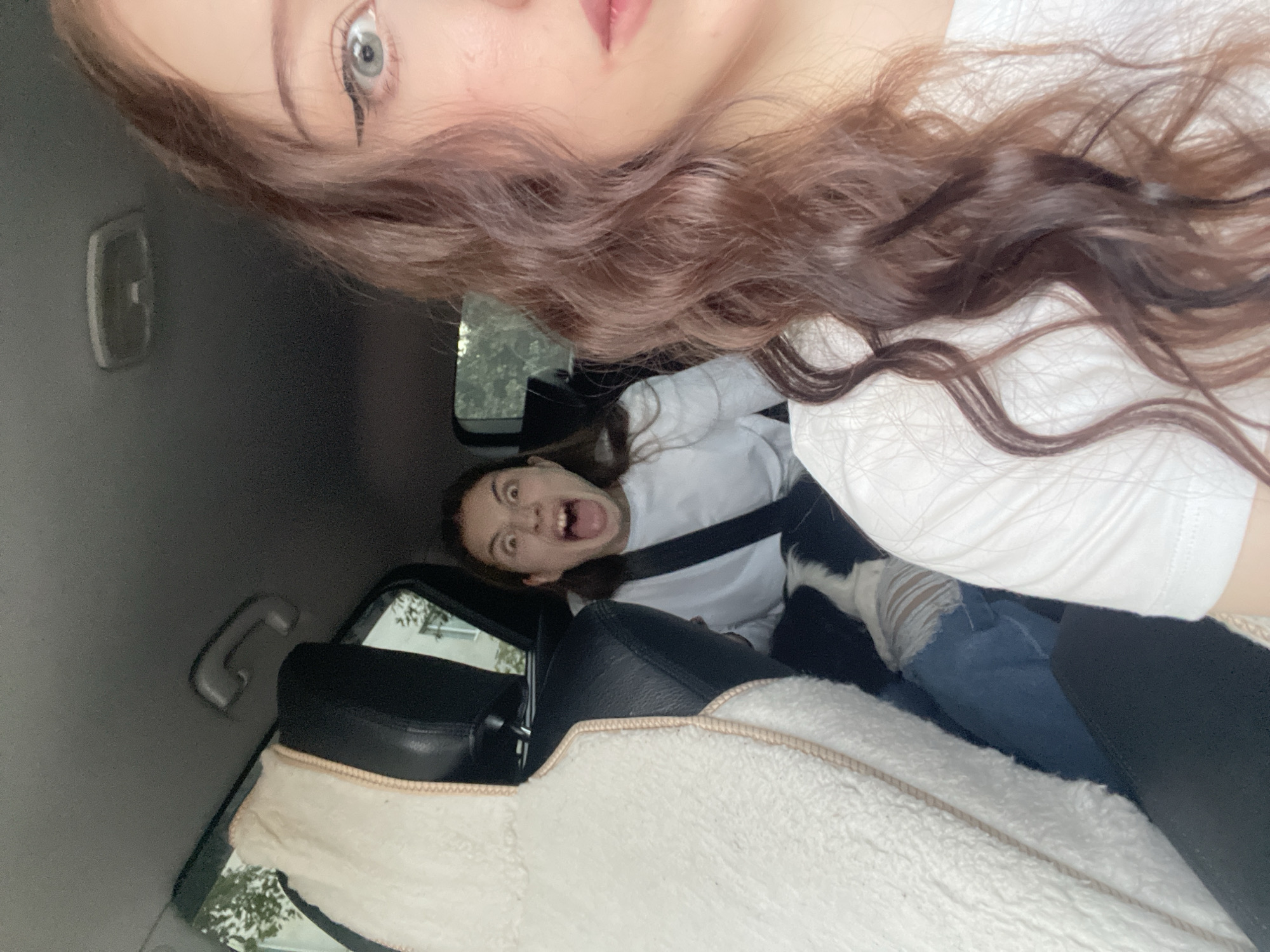 The image shows a young woman in the front passenger seat of a car, with her face close to the camera. Her brown hair is styled in loose waves, and she is wearing a white t-shirt. Her right eye is visible, with winged eyeliner. In the background, another young woman is in the back seat of the car, making a surprised or scared expression with her mouth wide open. She is wearing a white long-sleeved shirt and a seatbelt. A white fluffy car seat cover is visible in the foreground, obscuring part of the interior. The car appears to be in motion, as indicated by the slightly blurred background which shows trees and buildings, suggesting an urban or suburban environment. The lighting suggests it is daytime. The location is Votkinsk, Russia.