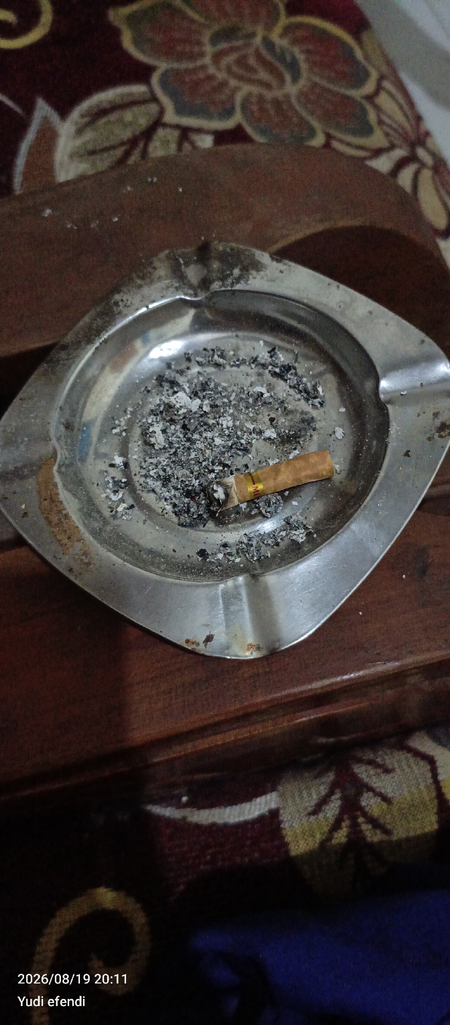 The image shows a close-up view of a used ashtray filled with cigarette butts and ash. The ashtray is made of metal and has a reflective surface. It is placed on a wooden surface, possibly a table, which is covered in part by a patterned fabric. The fabric has a floral design with colors of red, yellow, and green. The lighting in the image is dim, and there is a timestamp in the bottom left corner indicating "2026/08/19 20:11" and text that reads "Yudi efendi". The overall impression is that of a casual setting, potentially a home or a place where smoking is permitted. No specific details about the city of Rawajitu Selatan, Indonesia, can be inferred from the image itself.