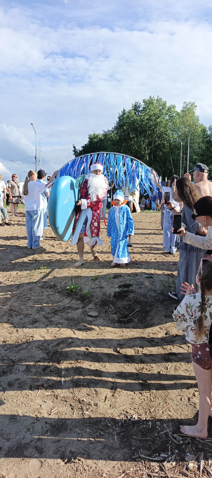 Two people dressed in costumes are walking on a sandy area. The person on the left is dressed as Santa Claus, complete with a red suit trimmed with white fur, a white beard, and a red hat. They are carrying a large, bright blue inflatable object, possibly a ring or a paddleboard. To the right of Santa is a child dressed in a blue costume with a hood, resembling a snow fairy or a winter spirit.
The scene appears to be outdoors on a beach or a sandy ground near a body of water, suggested by the presence of people in casual summer attire and the overall atmosphere. In the background, there's an arch decorated with numerous blue and white ribbons, adding a festive touch to the event. A crowd of people are gathered around, some observing the costumed individuals and others taking pictures with their phones. The time of day appears to be daytime, with clear skies and sunlight casting shadows on the ground, indicating fair weather. The general mood seems celebratory and lighthearted, possibly for a holiday event or a themed gathering, given the costumes. The location context is Chaikovsky, Russia. No visible text is present in the image.