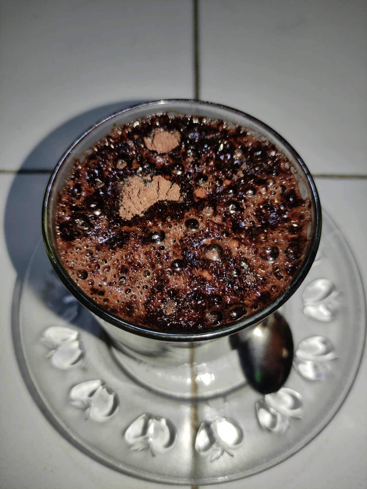 A close-up, top-down shot shows a glass of chocolate-colored beverage with a frothy top and a dusting of cocoa powder. The glass sits on a clear glass saucer with a decorative embossed pattern of leaves. A metal spoon rests on the saucer to the right of the glass. The background appears to be a tiled surface, with a shadow cast on the left side of the image. The scene is illuminated by a light source from the left, creating highlights and reflections on the beverage and spoon. The image appears to have been taken indoors, possibly in a home setting in Ngrendeng, Indonesia, given the presence of tiled surfaces often found in Indonesian homes.