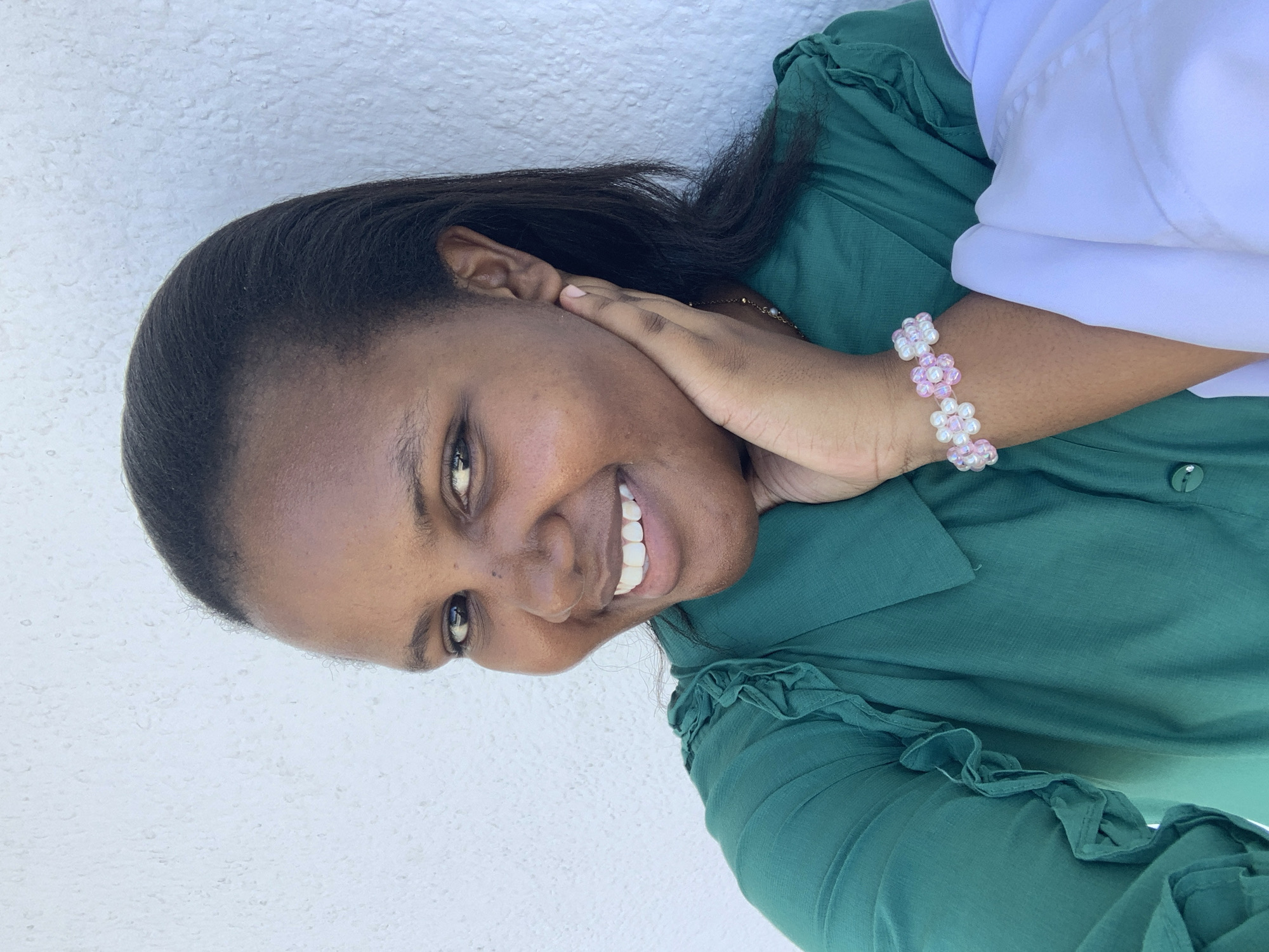 A woman is pictured in a close-up, smiling. She has dark hair, and is wearing a dark green button-up shirt with ruffles on the sleeves. Her hand, adorned with a pink and white flower beaded bracelet, rests on her cheek. A small necklace is visible. The background appears to be a white wall. The location of the image is unknown and there is nothing to suggest it was taken in Dar es-Salaam, Tanzania.