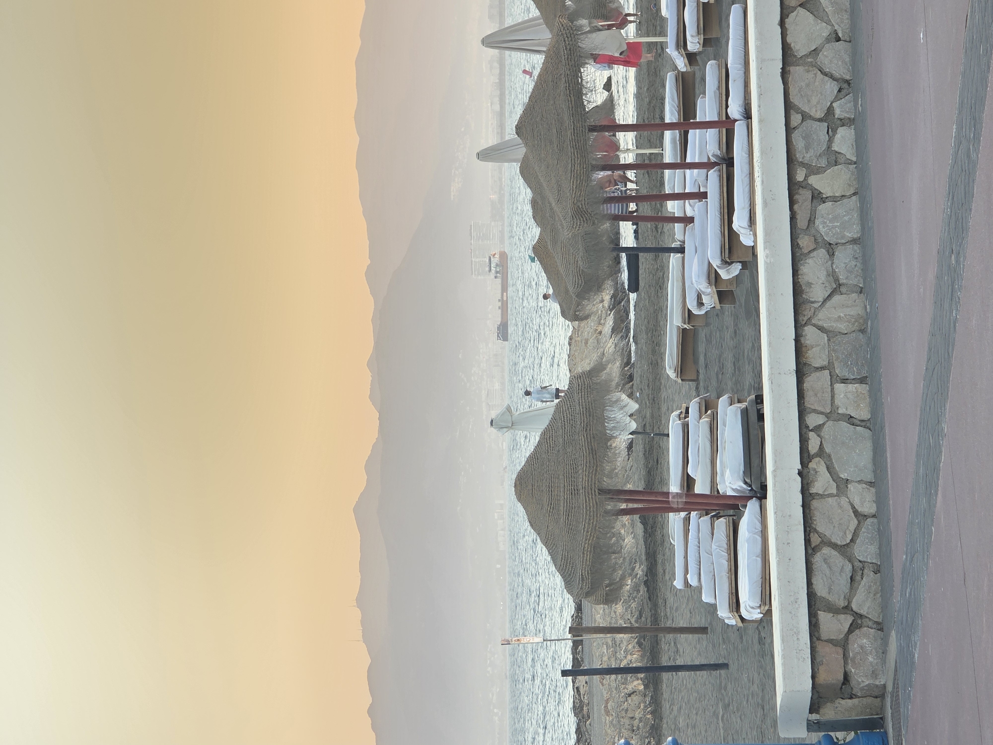The image depicts a beach scene in Málaga, Spain, likely during the late afternoon or early evening given the warm, hazy light and soft glow on the distant mountains. In the foreground, several rows of empty sun loungers are neatly arranged on the dark sand, covered with white sheets. Behind the loungers, thatched umbrellas provide shade. The sea is calm, with gentle waves lapping at the shore. In the middle ground, a few people are visible on the beach and in the water. A cargo ship is seen sailing on the horizon, and beyond it, the silhouette of mountains fades into the atmospheric haze. The overall impression is one of a peaceful, seaside resort.