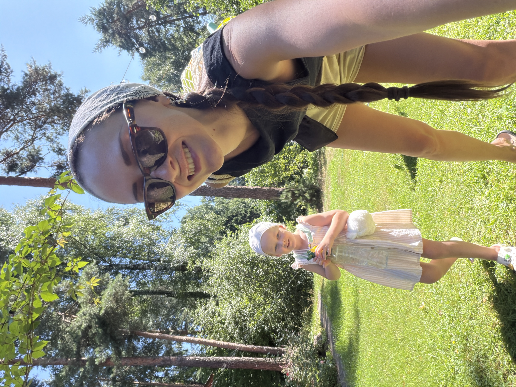 In a sunlit, grassy yard in Udelnaya, Russia, a woman with a bandana and sunglasses smiles into the camera, her long braid trailing down her back. She is wearing a black top and light-colored shorts. To her left, a young girl in a striped dress and a white headscarf holds a plastic bottle. The background is filled with lush green trees under a clear blue sky.