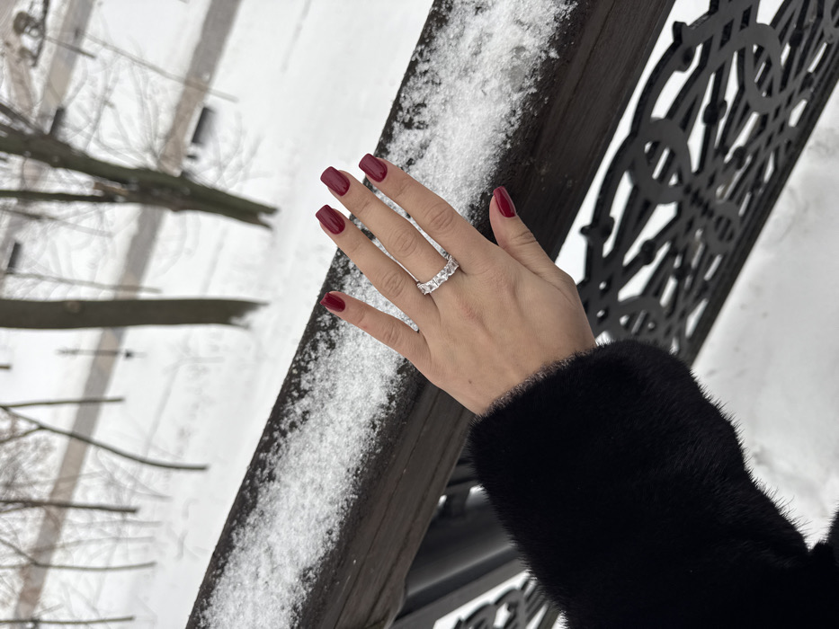 A close-up photograph displays an individual's left hand in a winter setting on the Pedestrian Bridge (Klitschko Bridge) in Kyiv, Ukraine, connecting Volodymyr Hill Park and Khreshchatyi Park. The hand, with fingernails painted dark red, wears a silver-colored ring featuring multiple rectangular-cut clear stones on the ring finger. The individual's black fur or faux-fur sleeve is visible at the lower left. The hand rests on a dark wooden handrail topped with fresh snow. Below the handrail is a dark, intricately patterned metal balustrade. The background shows a snow-covered park with bare trees and a visible path.