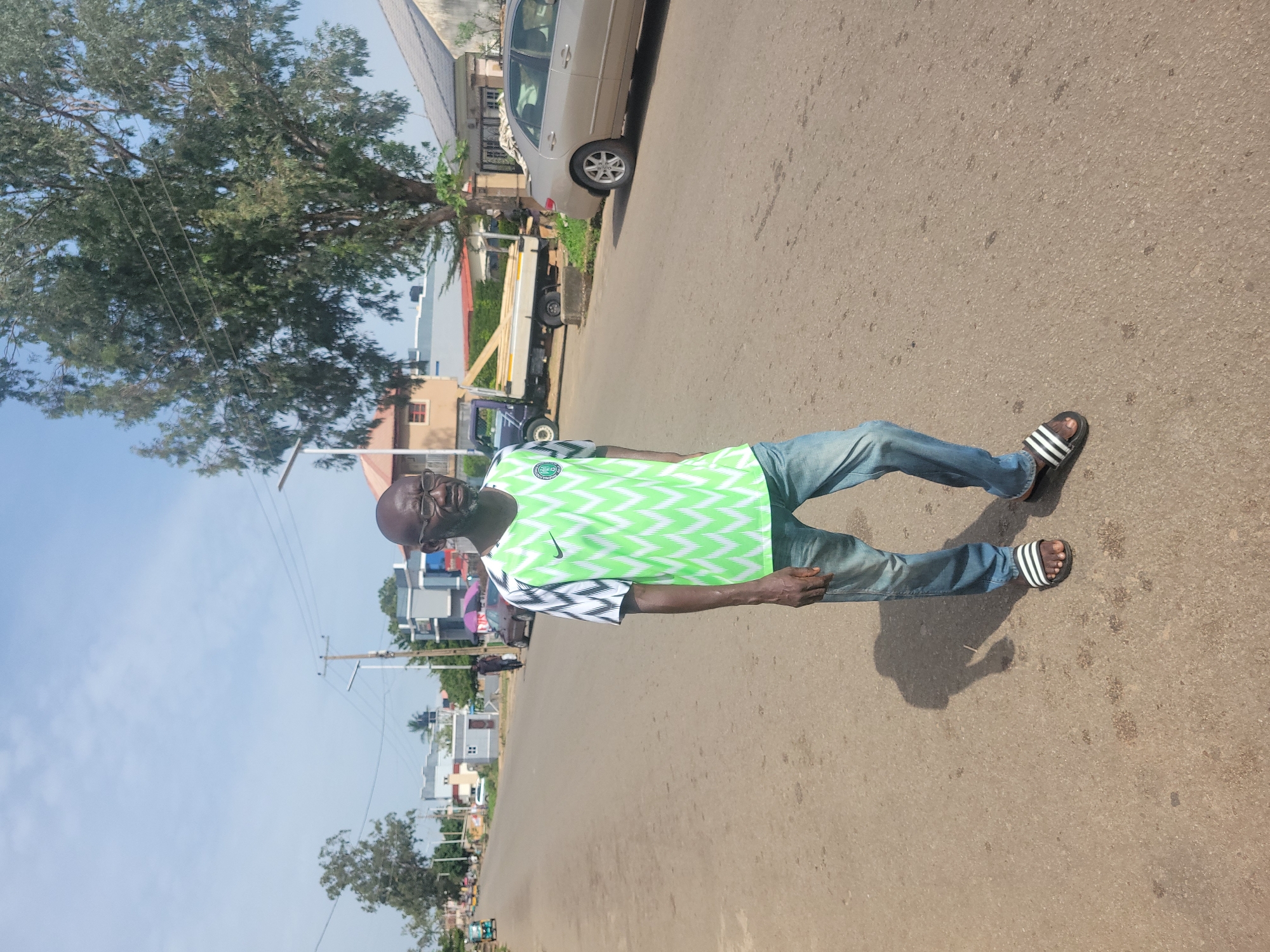 A middle-aged African man with a shaved head and glasses is standing on an asphalt road. He is wearing a green and white zigzag patterned jersey, blue jeans, and black and white striped slides. The man is looking to his right, with his body angled slightly to the left. His shadow is cast on the road to his left.
In the background, there is a light brown sedan parked on the right side of the road. Further down the road, there is a flatbed truck with lumber on its bed, and a person standing next to it. Several buildings are visible in the distance, including a two-story building with grey siding and a lighter colored building with a red roof. Trees line the right side of the road, with one large tree prominently in the foreground. Utility poles and wires are visible above the buildings and trees. The sky is blue with some scattered clouds. The scene appears to be in an urban or suburban setting in Jos, Nigeria.