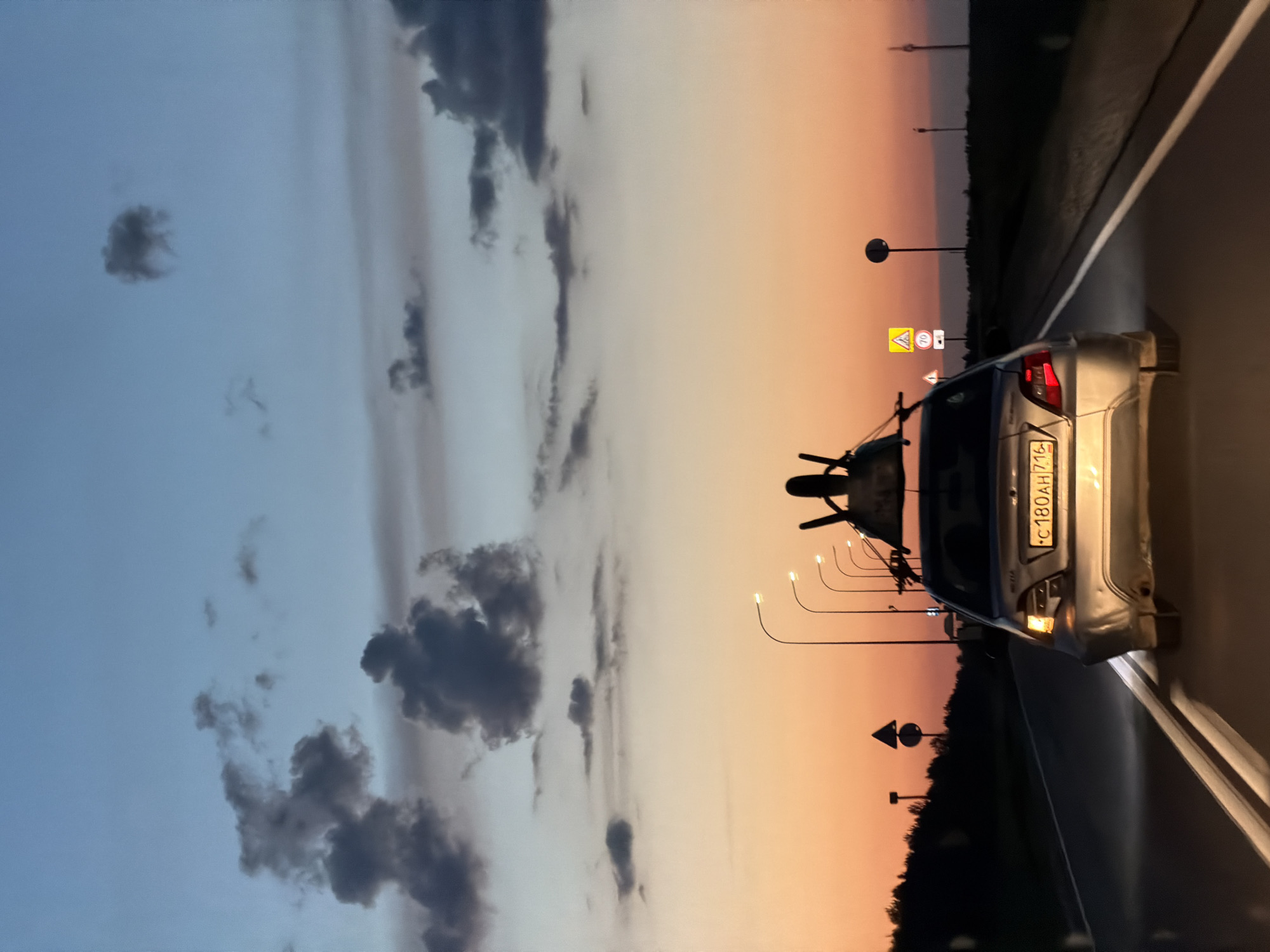 The image shows a silver sedan driving on a two-lane road at dusk. The car has a rack on its roof with some objects on it. The car's license plate is visible and reads "C 180 AH 716". There are streetlights visible along the side of the road, and various road signs, including a warning sign with a speed limit of 70. The sky is a gradient of blue and orange, with clouds. The environment appears to be rural or suburban, with a dark treeline visible in the distance. The overall impression is of a journey taken during the transition from day to night. The location is identified as Laishevo, Russia.